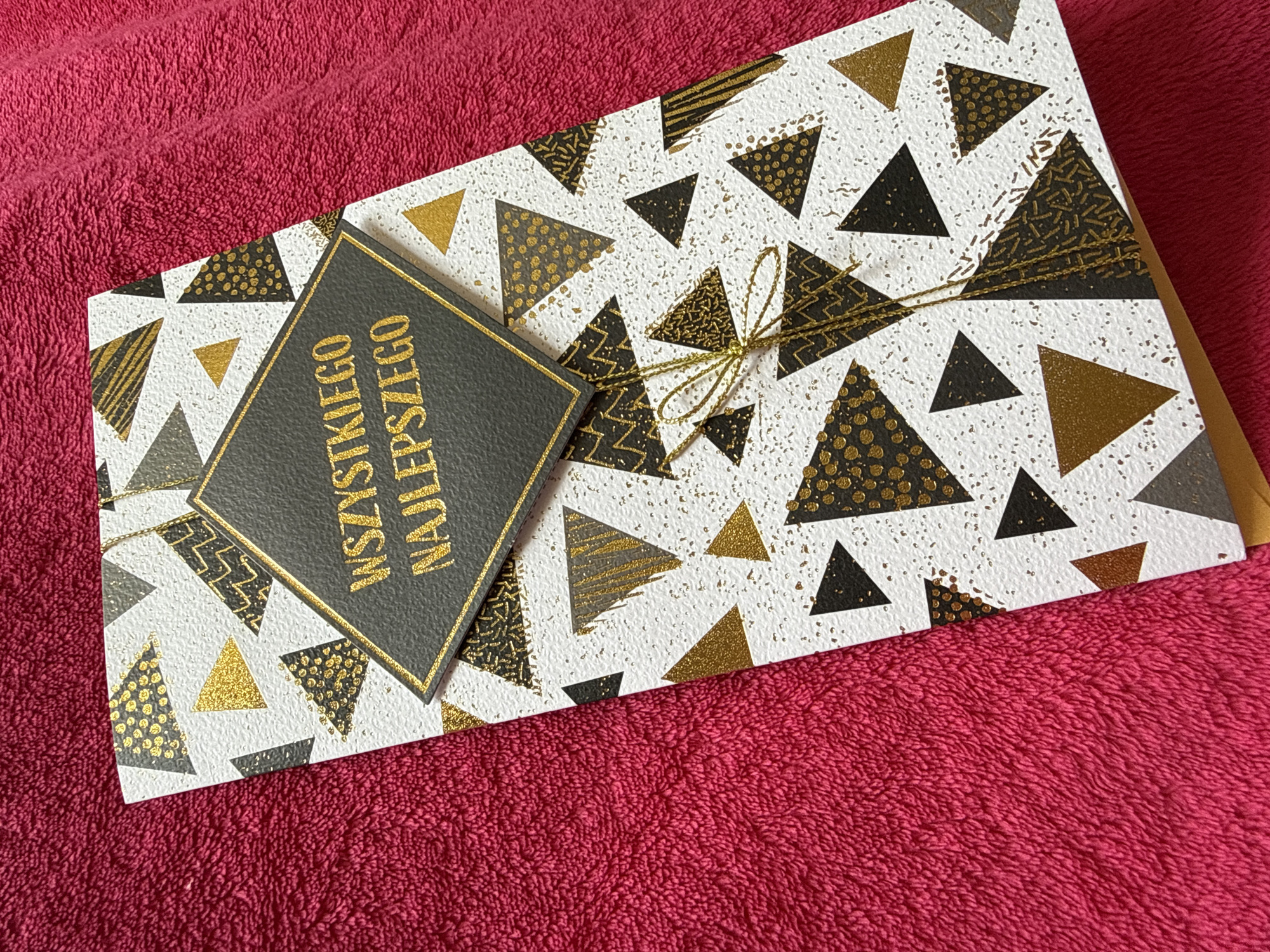 A greeting card with a geometric pattern is positioned slightly askew on a textured, magenta-colored fabric surface. The rectangular card features a white background flecked with gold speckles, adorned with numerous triangles. These triangles are rendered in solid gold, solid black, or black with gold-patterned infills such as dots, stripes, and abstract designs. A delicate gold string or twine is tied in a bow around the lower-middle section of the card. Overlying the upper-center of the card is a raised, diamond-shaped black plaque framed by a thin gold border. This plaque displays the gold-colored text "WSZYSTKIEGO NAJLEPSZEGO". The background fabric exhibits a dense, looped texture.