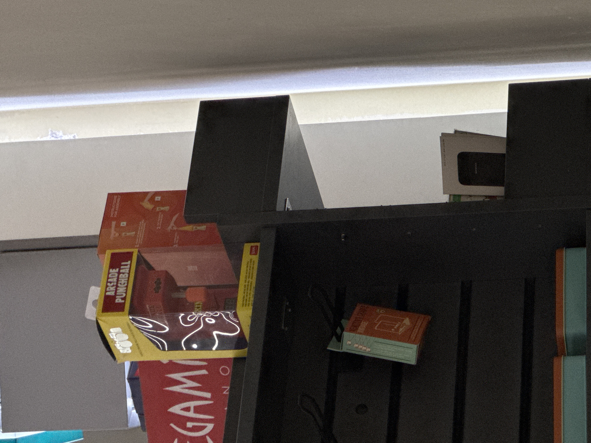 An image displays an indoor scene featuring a dark shelving unit against a light-colored background wall. The perspective is slightly angled, looking up towards the shelves.
On the left side of the shelving unit, a yellow box is prominently displayed, tilted forward. The box has text that reads "ARCADE PUNCHBALL" and features a "BOXER" logo. Its front surface is reflective with white swirling patterns. Partially visible beneath this box is a larger, reddish-orange box with the letters "MEGAM" visible, suggesting "MEGAGAMER" or similar. Further behind these boxes, an orange-red item with instructions is partially visible.
On the right side of the shelving unit, a stack of small, rectangular items (possibly mini notebooks or card decks) is arranged. These items have terracotta-orange tops and light blue-green spines. One piece of visible text on these items appears to say "PORTABLE" or "VINTAGE" and "GAME". Directly above and behind these items, a dark object, possibly a smartphone or small electronic device, is partially visible within a white box.
The shelving unit itself is dark, possibly black or dark grey, with vertical slats visible on the right side. Above the shelving unit, a bright, horizontal light source or ceiling edge illuminates the top of the scene, casting a glow on the light-colored wall or ceiling above. No people are present, and no activity is depicted beyond the arrangement of objects. The scene appears to be static, possibly a retail display or storage area.