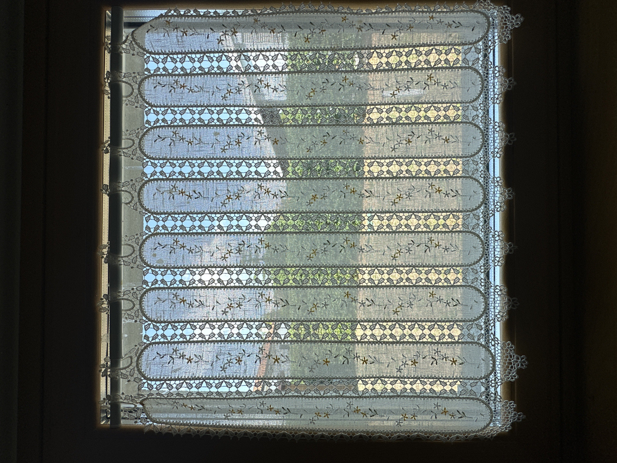 The image shows a close-up of a white lace curtain hanging in a window. The curtain has a scalloped bottom edge with intricate floral patterns. Vertical bands of lace with diamond-shaped cutouts run throughout the curtain, interspersed with embroidered floral sprigs. Through the semi-transparent fabric of the curtain, a view of green foliage and a house with a terracotta roof is visible. The scene appears to be set during the daytime, with natural light illuminating the interior through the window. The overall impression is one of a domestic interior with a view of an outdoor garden. The location of the image in Trieste, Italy, is not directly discernible from the visual elements alone, as the architecture and landscape are not distinctly identifiable as belonging to that specific city without external information.