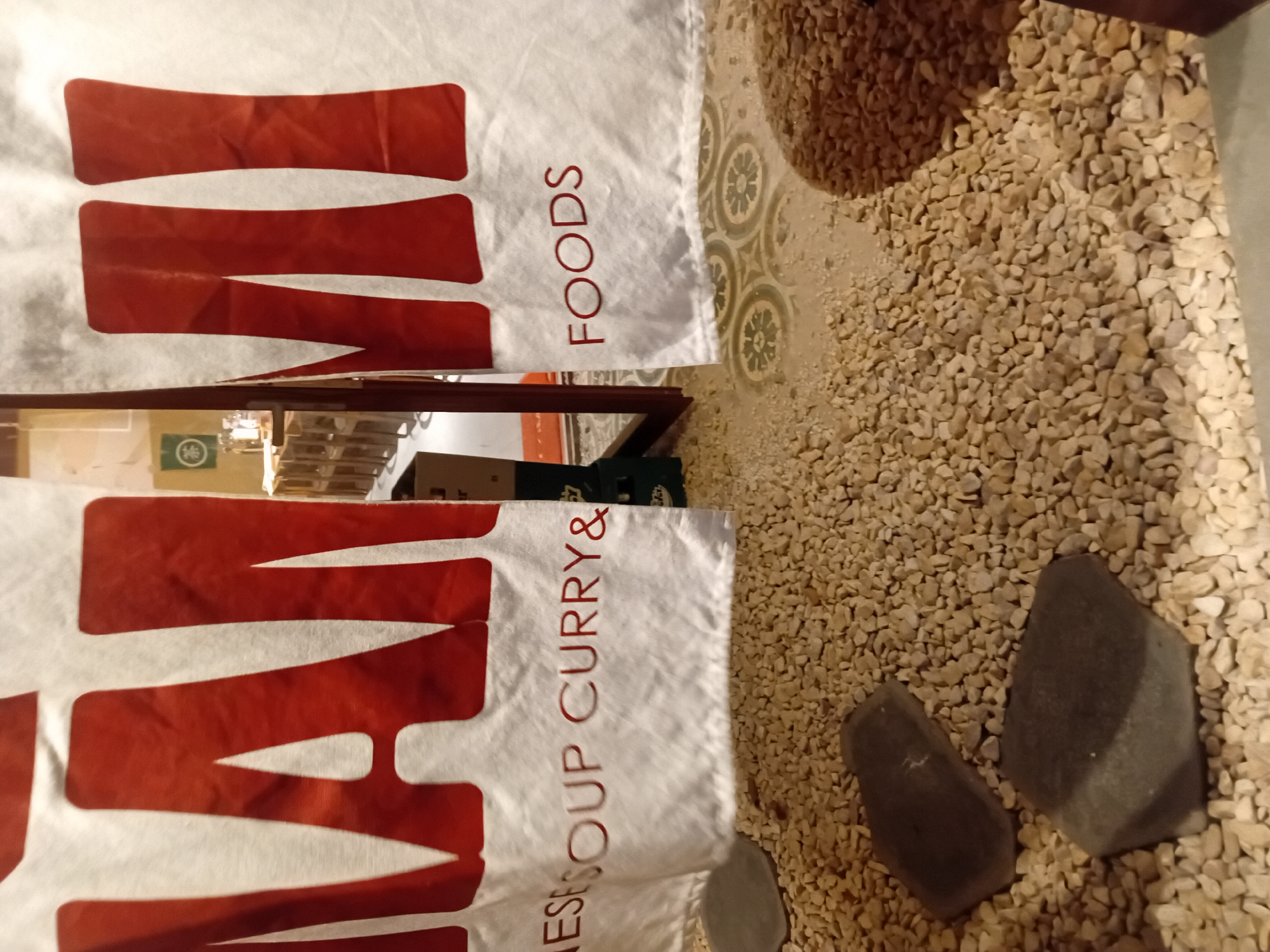 The image captures a close-up view of a white curtain with large red block letters that spell "MANMI" across the top. Below the large letters, smaller red text reads "ESE SOUP CURRY & FOODS". The curtain is partially obscuring an indoor scene. The foreground is covered in small, light-colored pebbles, with two dark, flat stones positioned on them. In the background, through a doorway or opening in the curtain, a dining area with wooden tables and chairs is visible, along with a green banner featuring a circular symbol. The flooring in the background appears to be tiled with a decorative pattern. The overall impression is that of an entrance to a restaurant, possibly a Japanese cuisine establishment, given the text on the curtain. The location is not specified, but the requested information about Đà Nẵng, Vietnam is not present in the image itself.