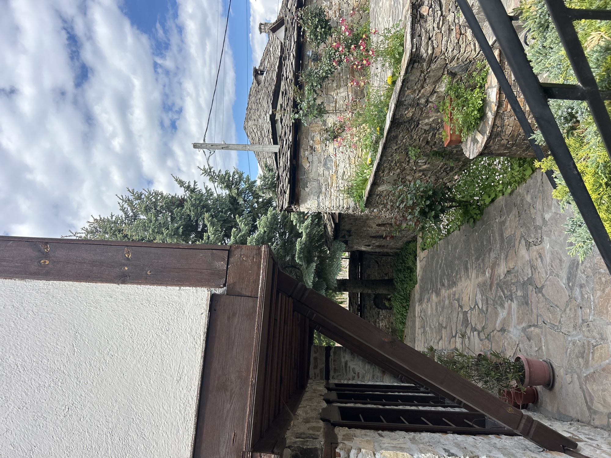 This image captures a view of a narrow passageway in Dolen, Bulgaria. On the left is a white building with dark wooden trim and a roof overhang. To the right, a stone wall is adorned with plants and flowers. The passageway is paved with stone and leads toward a structure with a slate roof. Overhead, a cloudy sky is visible. The image is slightly rotated counterclockwise.