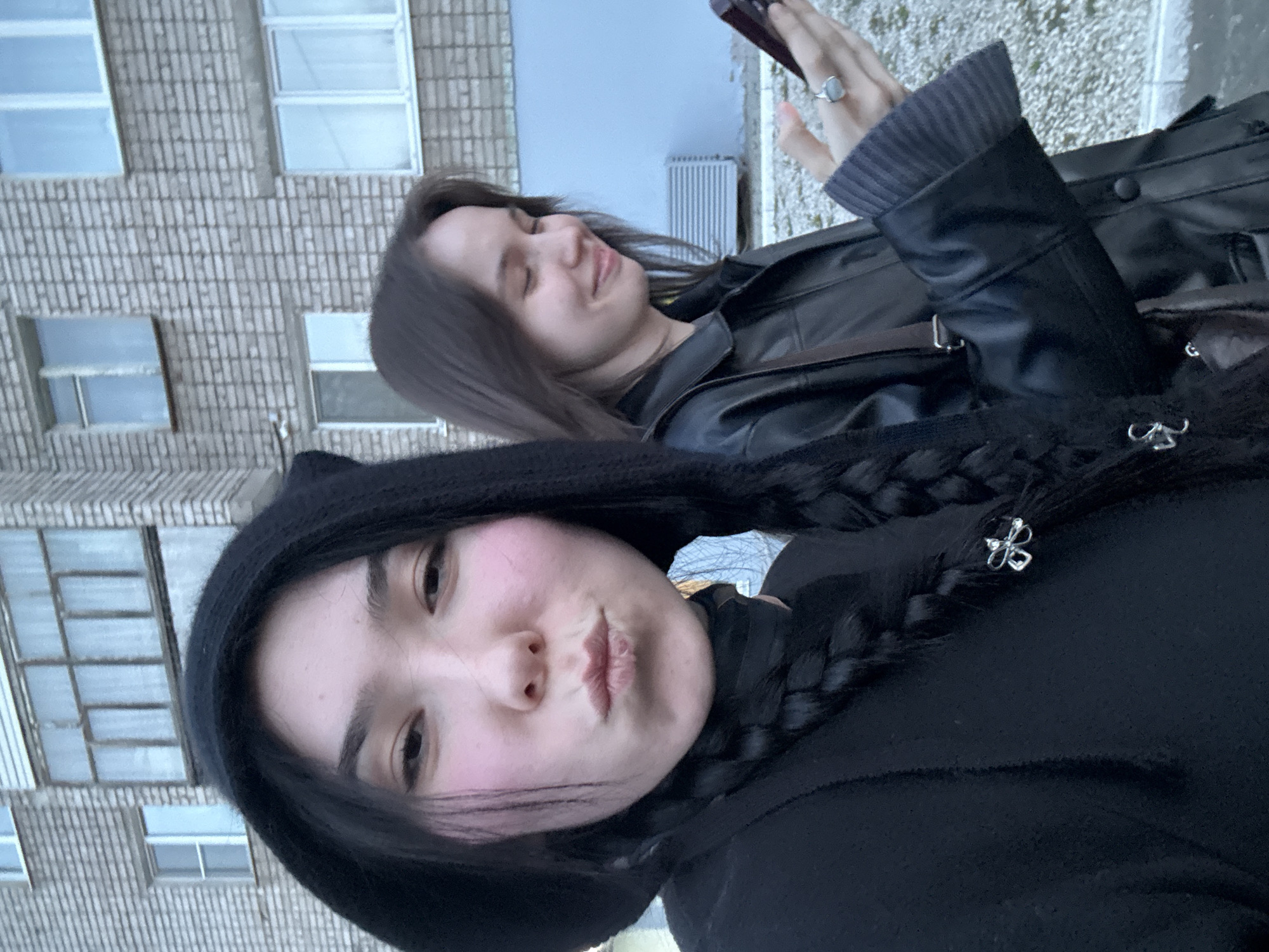 A selfie captures two young women outdoors in Izbyshev, Russia. The image is oriented with the subjects and background tilted to the right. In the foreground, a young woman with dark hair, wearing a dark hoodie, makes a playful pouty face directly at the camera. Her cheeks are visibly flushed with pink blush, and her long dark hair is styled in braids adorned with small silver butterfly-shaped clips.
Slightly behind and to her right, another young woman with dark hair and a black leather jacket smiles softly with her eyes closed or squinted. She is holding a dark-colored smartphone in her right hand, and a silver ring is visible on her left index finger.
The background features a multi-story building constructed of light-colored brick, characterized by numerous rectangular windows that reflect the overcast sky, indicating daytime. A metallic, finned object, possibly a heating unit or architectural detail, is visible on the ground level behind the second woman. The overall impression is of a casual, friendly moment captured in an urban setting during cool weather, likely autumn or spring, given their attire. There is no visible text in the image.