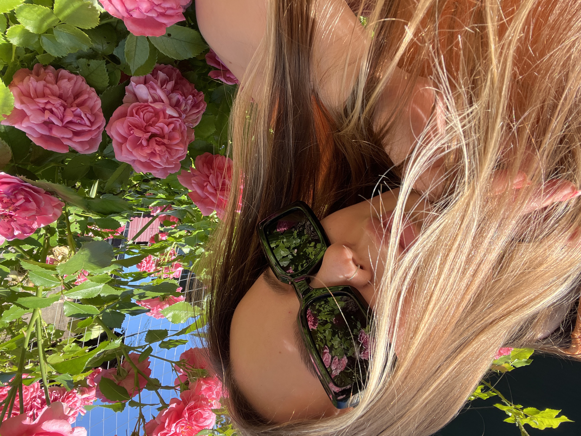 The image shows a close-up of a young woman with long blonde hair, wearing sunglasses. The sunglasses reflect pink roses and green foliage. She is outdoors, with a rose bush prominently featured in the foreground and background. The sky is visible and appears blue. The overall impression is of a sunny day. The location is identified as Novopokrovskaya, Russia.