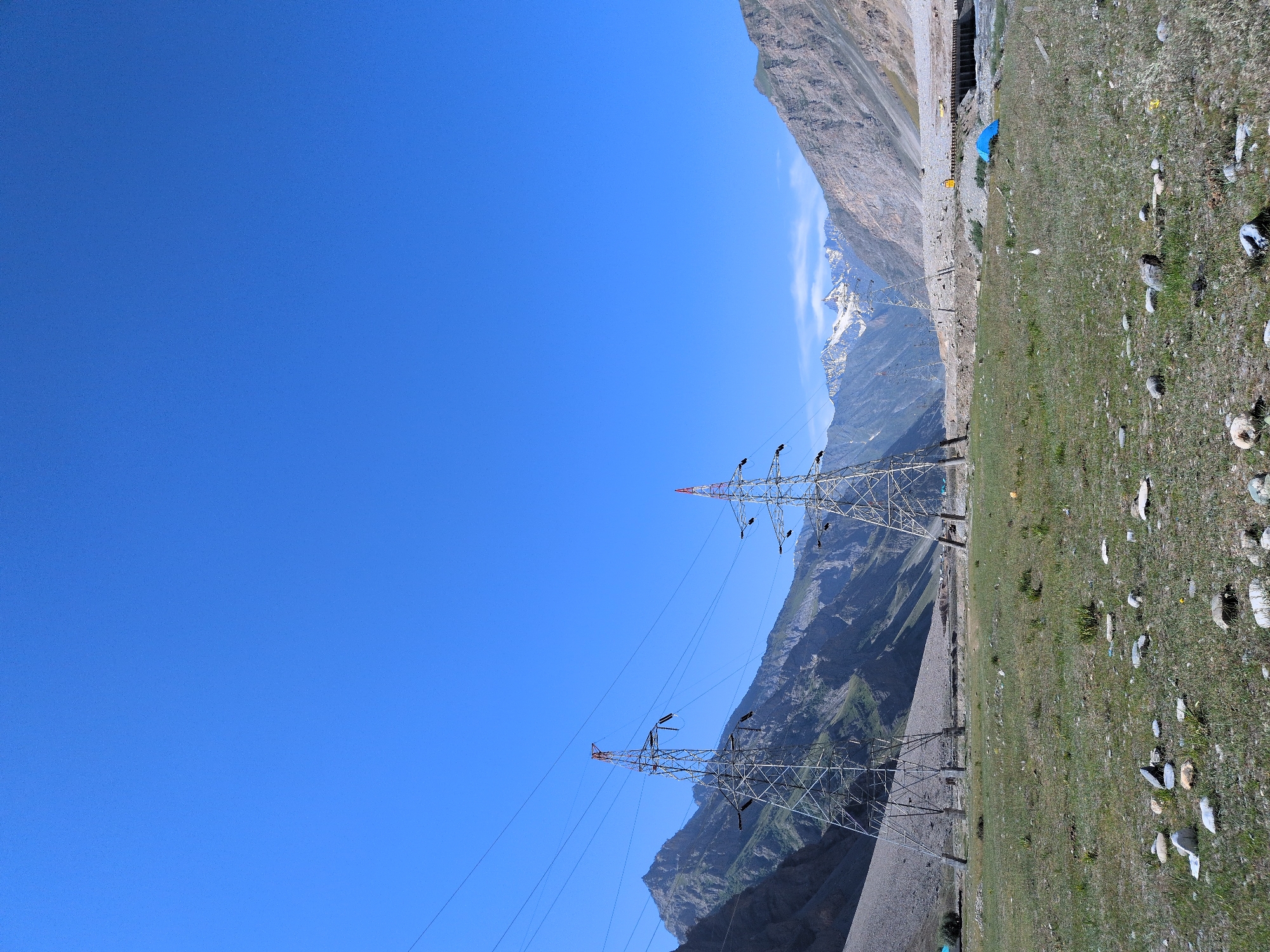 This is a landscape photograph taken in Matayeen, India, under a clear blue sky. The foreground features a grassy field scattered with rocks. Two large, latticed metal electricity pylons stand prominently in the mid-ground, with power lines extending between them and into the distance. Beyond the pylons, a valley unfolds with steep, rocky mountains on either side, some of which are snow-capped in the distance. The terrain is rugged and sparsely vegetated. In the far mid-ground, a blue tent is visible on the right side of the frame, near what appears to be a body of water or a riverbed and a bridge. The overall impression is one of a remote, mountainous region with signs of infrastructure.