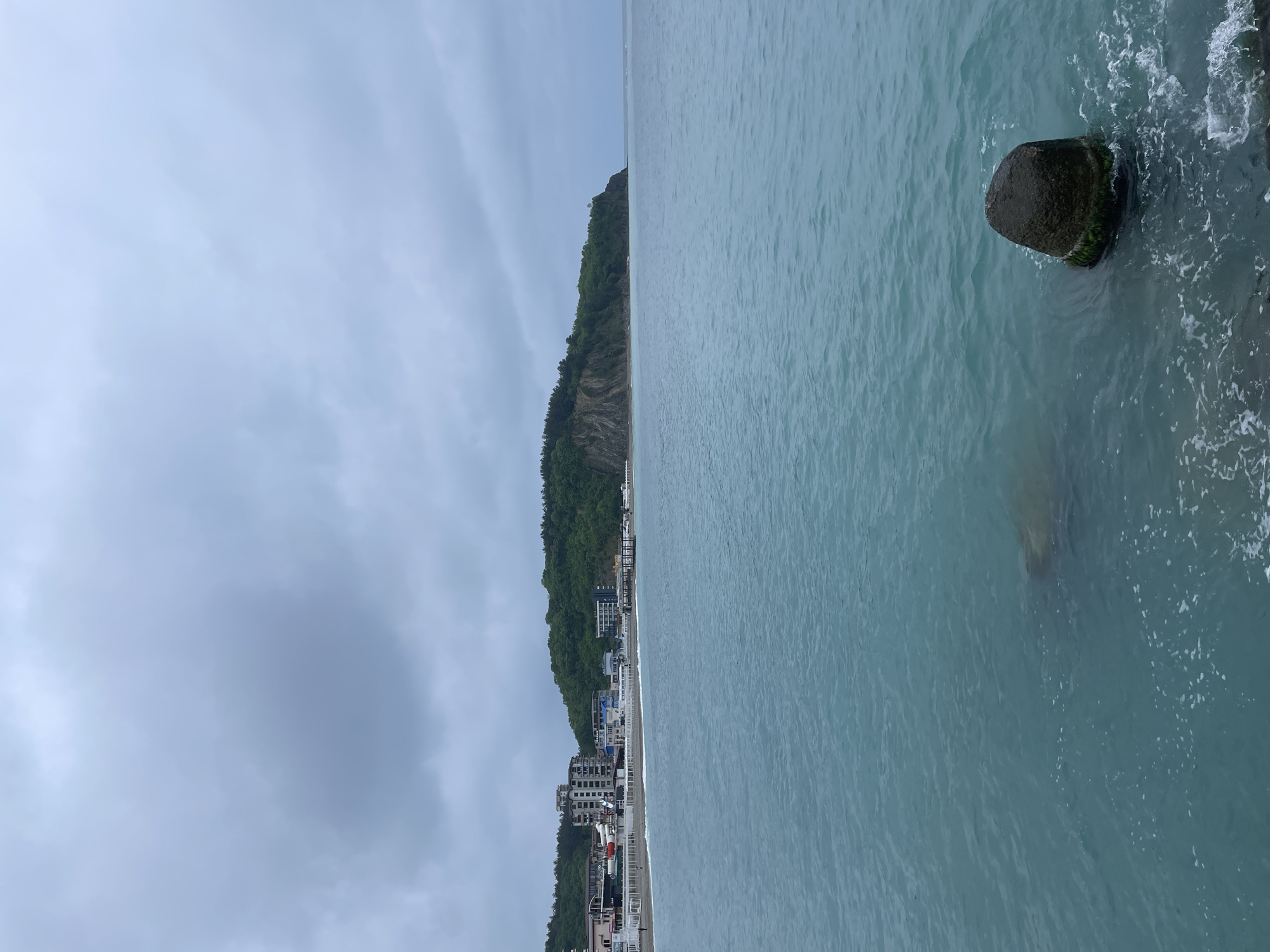 The image shows a view of the Black Sea coast in Olginka, Russia. The sea is a light turquoise color with slight waves. A large, moss-covered boulder sits in the water close to the right edge of the image. The shoreline, visible on the left, features a row of buildings extending along the coast. A steep, tree-covered cliff rises behind the buildings. The sky is overcast with a cloudy, grey expanse covering most of the upper part of the frame. The photograph appears to be taken from an elevated position.