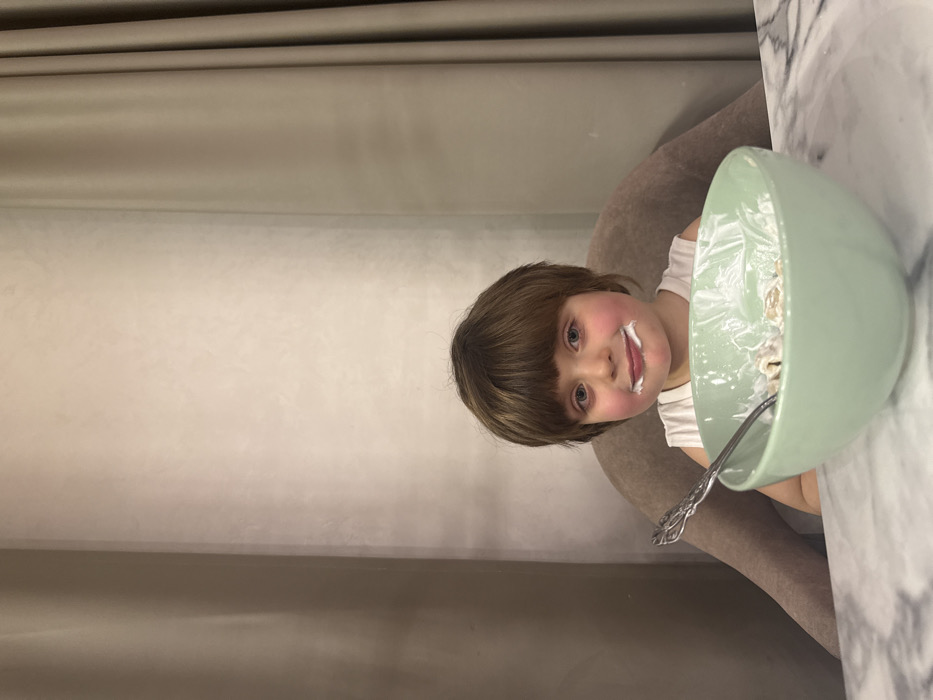 A young child with brown hair and rosy cheeks sits at a table with a large, light green bowl filled with cereal and milk. Some milk is smeared on the child's face. A silver spoon rests in the bowl. The table has a marble-like surface. Behind the child is a light brown, padded chair and a neutral-toned wall. The photo has been rotated, so the scene is oriented sideways.