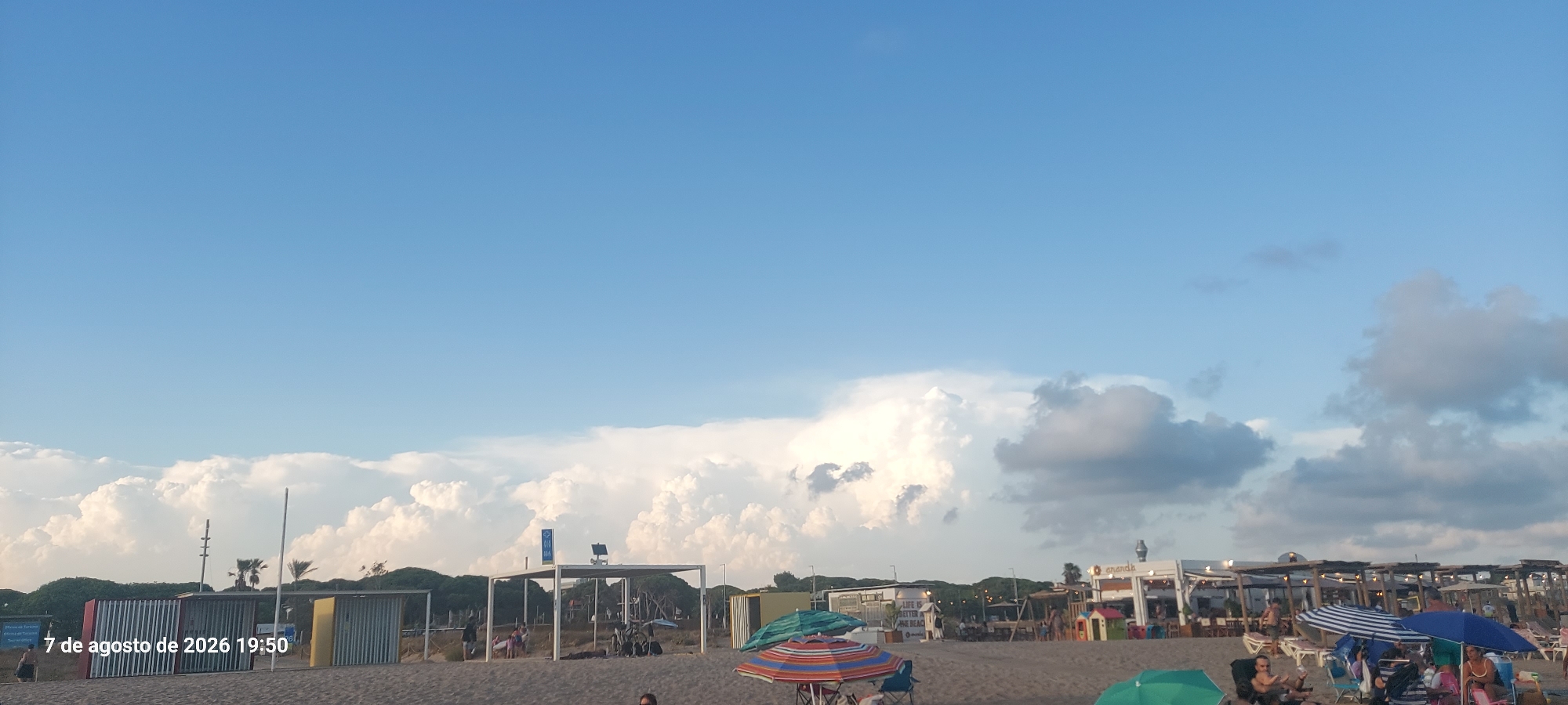 The image depicts a beach scene in Castelldefels, Spain, on August 7, 2026, at 19:50. The sky is a clear blue with scattered cumulus clouds, some of which are large and fluffy, illuminated by the setting sun. The foreground consists of a sandy beach with several beach umbrellas, chairs, and people. In the middle ground, there are structures that appear to be beach facilities, possibly changing rooms or small kiosks, with corrugated metal walls. Some of these structures have awnings or roofs supported by metal frames. Further back, a line of trees forms a backdrop to the beach facilities. A white building with a sign that says "ananda" and other smaller structures are visible to the right, suggesting a commercial area or beach club. People are seated in chairs, some under umbrellas, and others are walking or standing on the sand. The overall impression is that of a late afternoon on a crowded beach, with the sun beginning to descend.