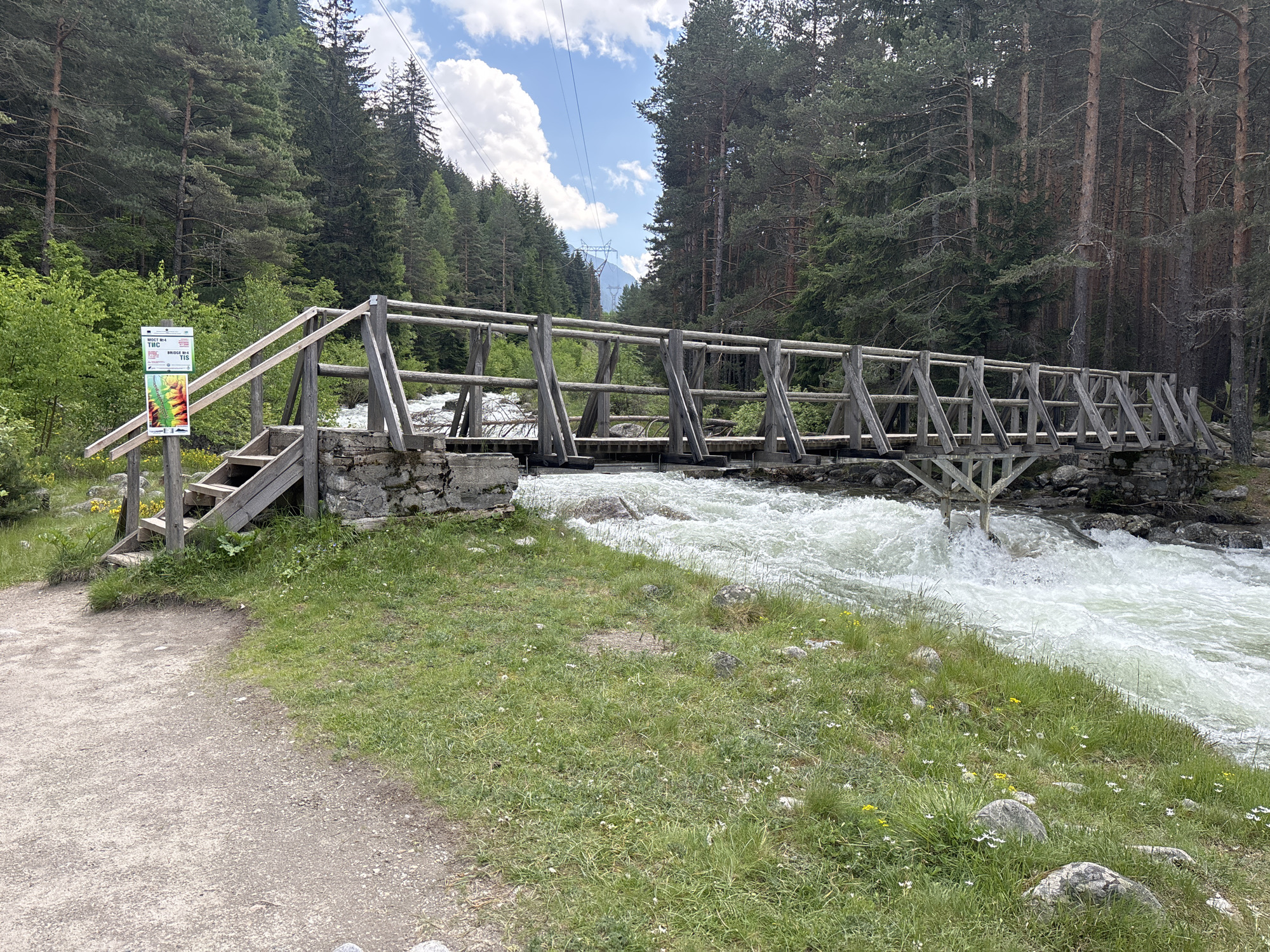 A long shot captures a weathered wooden bridge spanning a turbulent, white-foamed river in Beli Iskar, Bulgaria. The bridge, constructed from rough-hewn logs and planks, features a series of angled, cross-braced railings and a flat deck. Its near end is supported by a stone and concrete abutment, from which a short set of wooden steps descends to a dirt path. The river flows rapidly from left to right, its surface agitated by rocks and a strong current.

On the left side of the frame, near the bridge's entrance, a rectangular information sign is affixed to a wooden post. The sign has a white upper section with black text, including "МОСТ №4" (Bridge No. 4) and "BRUGGE №4," and a lower section displaying a colorful topographical map. The dirt path occupies the lower-left corner, transitioning into green grass with scattered white and yellow wildflowers along the riverbank.

Dense coniferous forests, dominated by tall, dark green pine trees, line both sides of the river and extend up the steep mountain slopes, framing the scene. Above the treeline, a partially visible blue sky with scattered white cumulus clouds suggests fair weather. High-tension power lines traverse the sky, stretching across the valley, with a distant pylon faintly discernible. No individuals or animals are present in the scene.