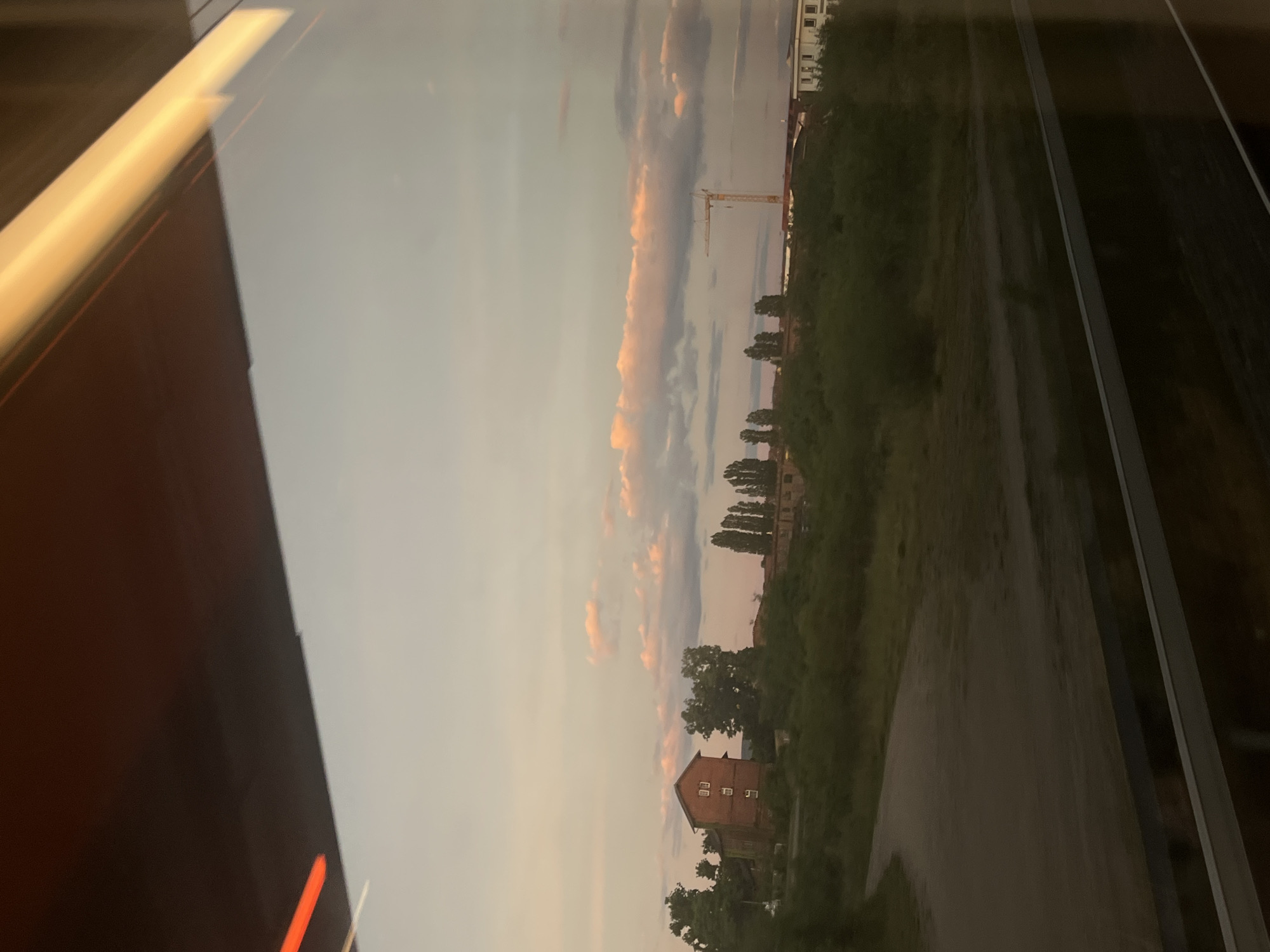 An outdoor, eye-level, medium shot captures a view through a train window. The scene depicts a rural landscape at dusk. In the foreground, the window frame is visible, with reflections of lights and the interior of the train. The middle ground shows a winding dirt road and a grassy field with a cluster of trees and a brick building. In the background, more trees and buildings are visible, along with a construction crane. The sky is a soft gradient of pink, orange, and gray, with scattered clouds. The overall impression is one of serene natural beauty, tinged with the signs of human activity.