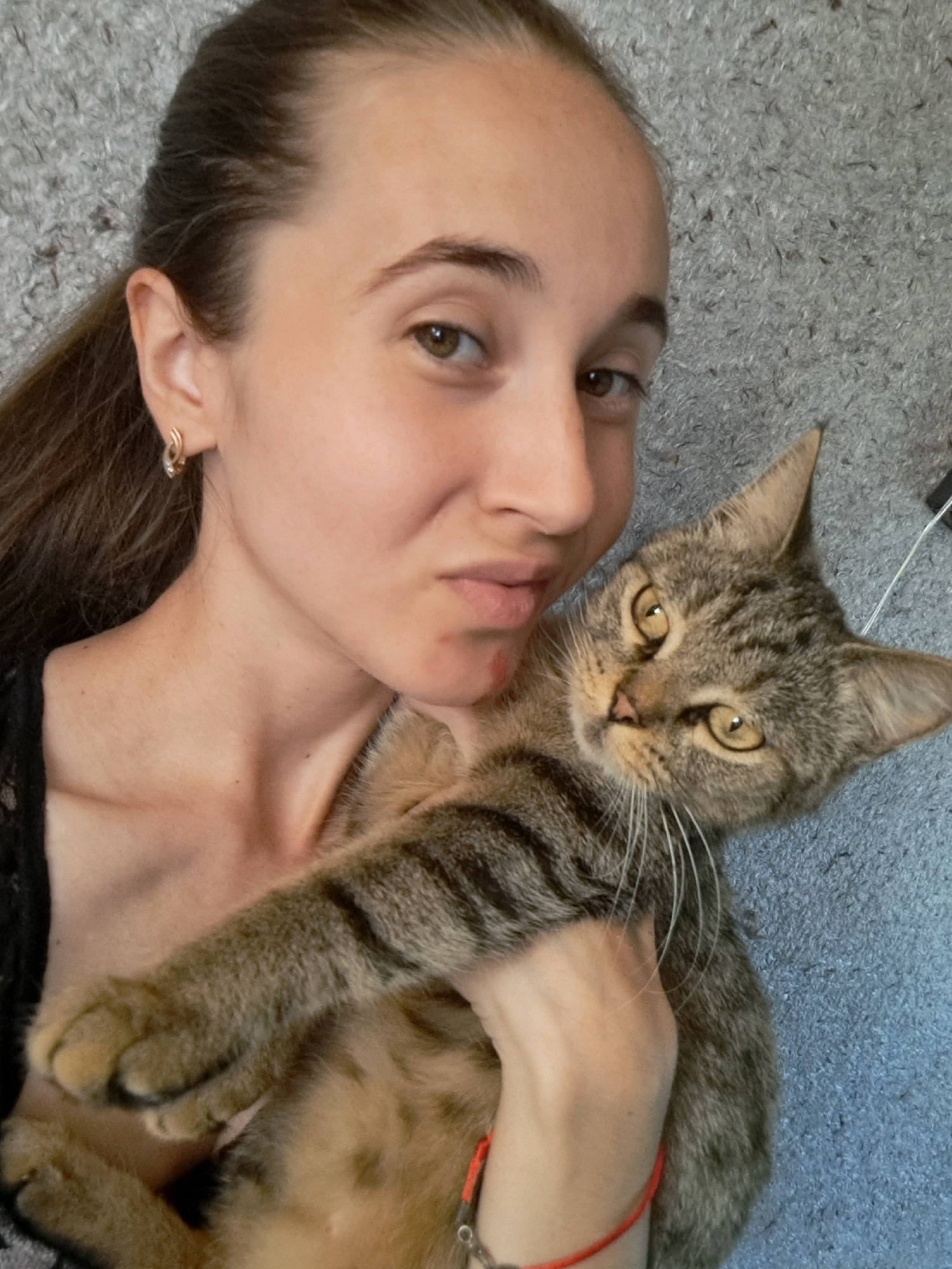 A young woman with brown hair and brown eyes is taking a selfie with her cat. She is wearing a black top and gold earrings. The cat is a tabby with green eyes and is being held in her arms. The background is a gray, textured surface, possibly a carpet or wall. The image is taken indoors. There is no indication that this image is located in Vypasne, Ukraine.