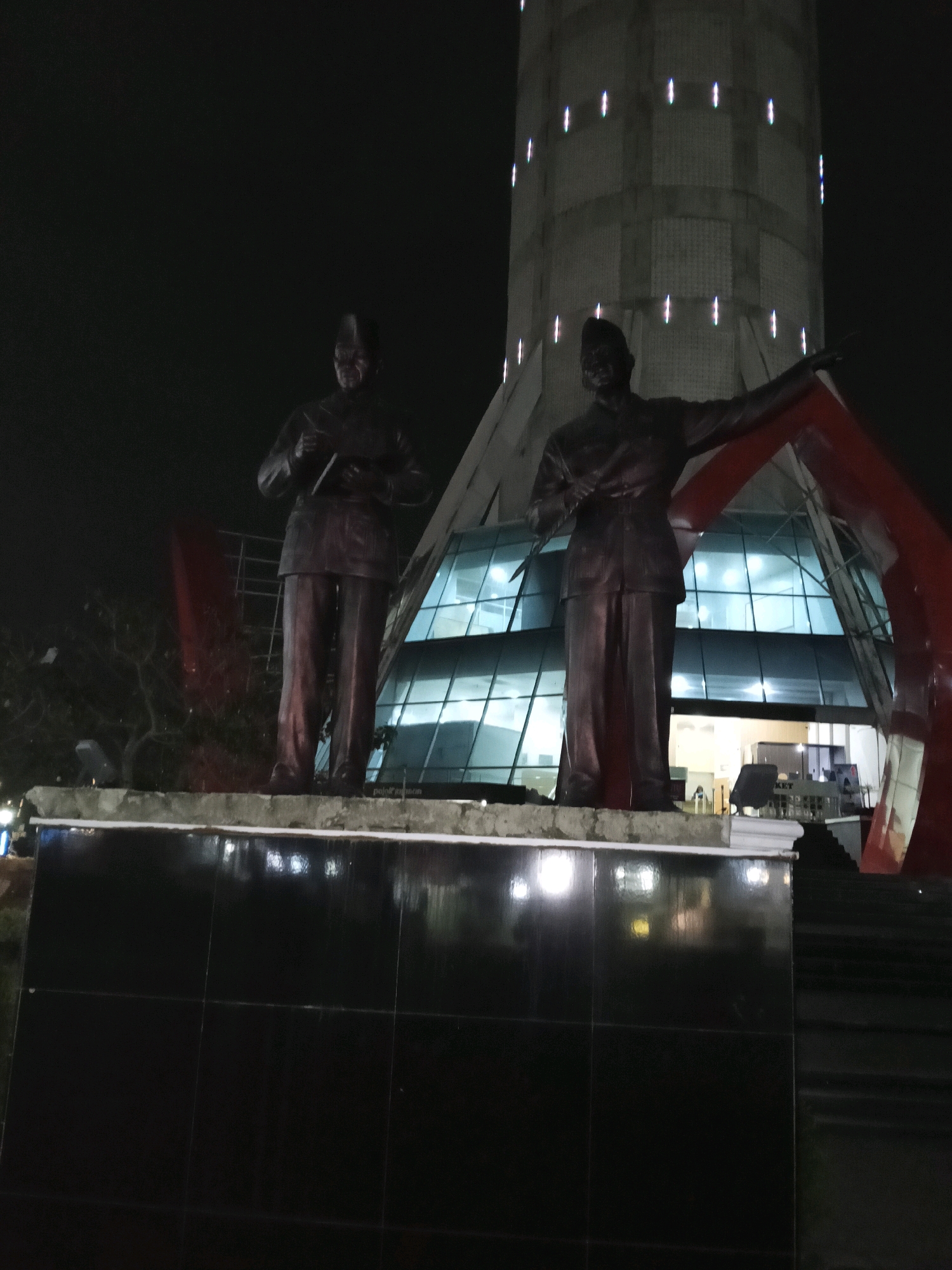 The image displays a night-time view of the Satria Gatotkaca Monument in Purwokerto, Indonesia. Two dark bronze statues of men stand on a black tiled platform in the foreground. The statue on the left holds an object in its hands, while the statue on the right points with its right arm and holds an object in its left hand. Behind the statues, a modern building with a glass facade and a cylindrical tower are visible. The building is illuminated from within, and the tower is adorned with vertical white lights. Red angular structures frame the building and extend towards the statues. The overall scene is dark, with reflections from the building and lights visible on the wet surface of the platform.
