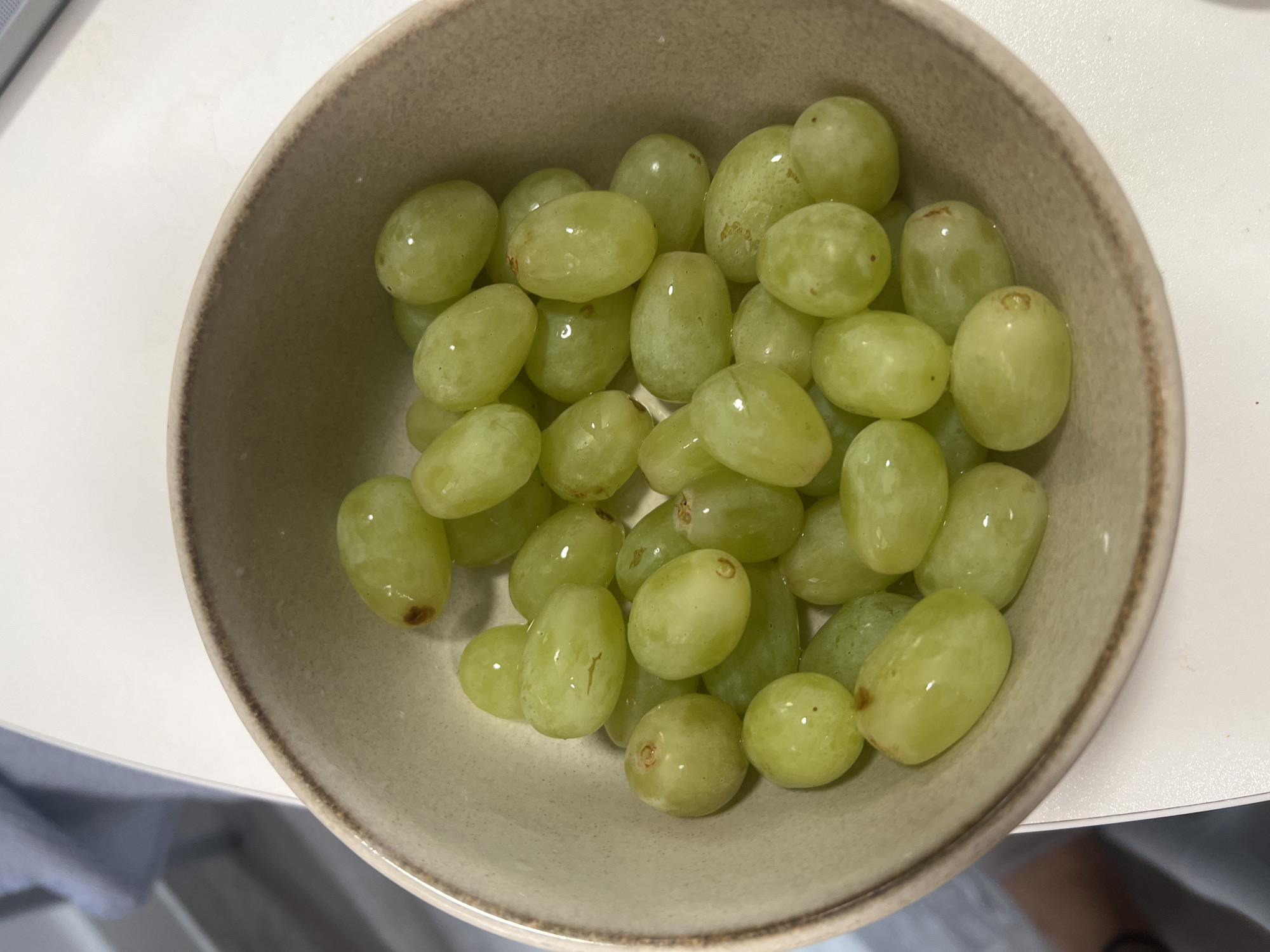 A top-down, eye-level shot shows a bowl of green grapes. The bowl is light brown with a dark brown rim, placed on a white surface. The grapes are mostly oval in shape, densely packed in the bowl, and light green, some with slight variations in color and texture. The background is out of focus, showing glimpses of white and gray. There is no evidence to pinpoint the image's location in Epsom and Ewell, United Kingdom.