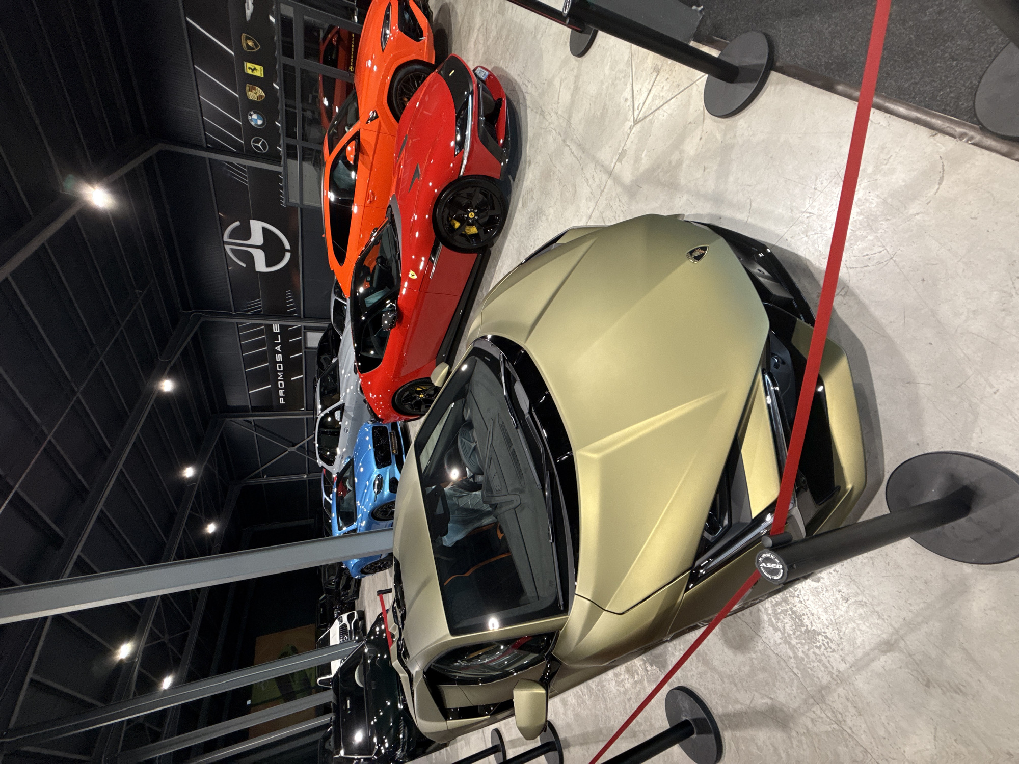 A high-angle, eye-level full shot shows a gold Lamborghini Aventador parked in the foreground of a dealership showroom in Plovdiv, Bulgaria. The car is angled slightly to the right, with its front end facing the viewer. The car's interior, including black and orange seats, is visible through the windshield. A red velvet rope is strung across the front of the Lamborghini, supported by two black stanchions.
Behind the Lamborghini, a red Ferrari Roma is parked, its front end also angled to the right. The Ferrari's bright red paint contrasts with the gold of the Lamborghini. Further back, an orange Lamborghini Urus SUV is visible, parked parallel to the Ferrari. In the background, several other luxury cars, including a blue Bentley, a white BMW SUV, and a black sedan, are parked. The showroom has a dark, industrial ceiling with exposed beams and track lighting. The walls feature large banners with logos and branding, including what appears to be a stylized logo on a black banner with "PROMOSALE" text. The floor is a polished concrete surface. No people are directly interacting with the vehicles, suggesting a static display.