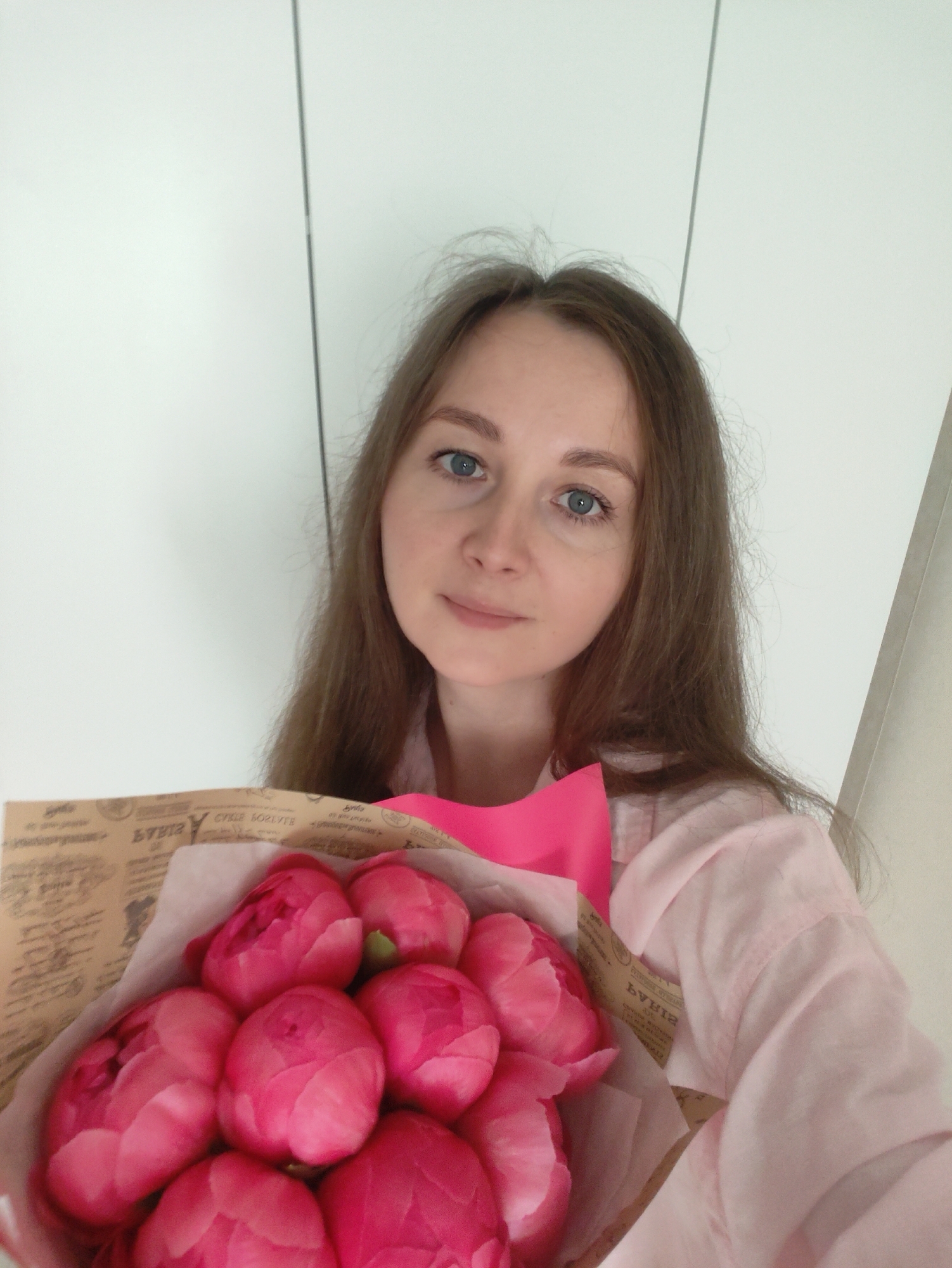 A close-up, selfie-style portrait captures a woman holding a large bouquet of pink peonies. The woman, with long brown hair and blue eyes, looks directly at the camera with a neutral to slightly smiling expression. She is wearing a light pink, long-sleeved shirt.
The bouquet prominently features numerous vibrant pink peonies, most of which are in tight bud form. They are wrapped in brown paper that has stylized, inverted text and patterns, resembling vintage newspaper print. Visible text fragments on the wrapping paper include "БИБАЯ" (BIBAYA) and "DYEIH", which appear to be decorative. A bright pink ribbon or trim accents the top of the bouquet's wrapping.
The background is a plain, light-colored (likely white or off-white) indoor wall, possibly a closet or cabinet door, indicated by a faint vertical seam. The lighting suggests an indoor setting, likely during the daytime or in a well-lit interior. The image conveys a casual, pleasant moment, possibly celebrating the reception of the flowers.
