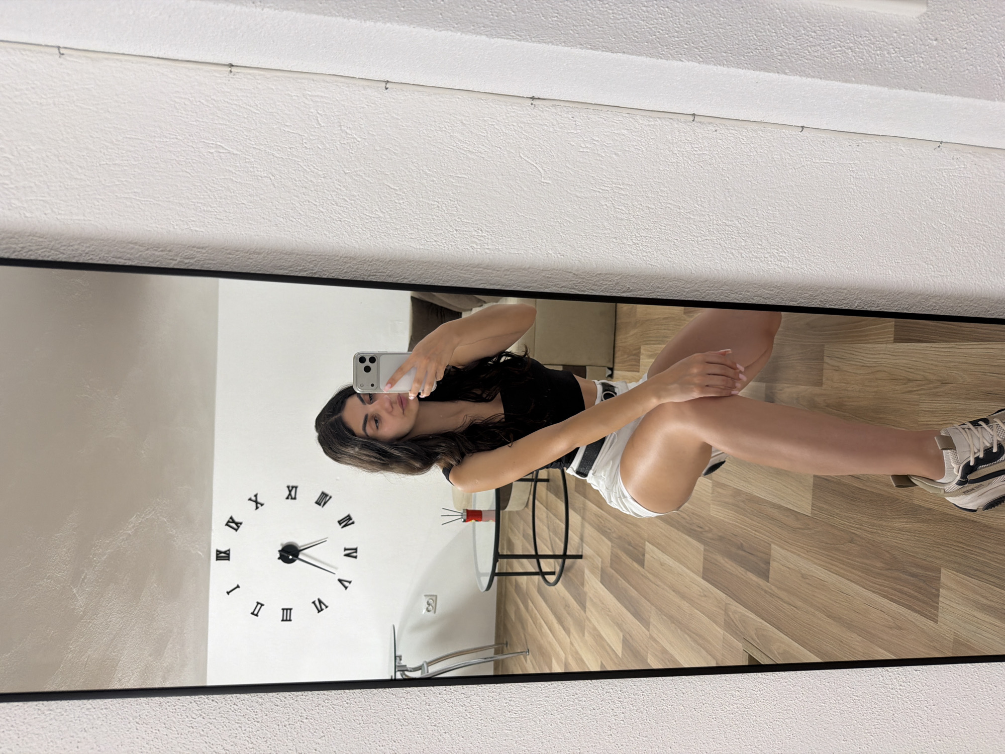 This is a mirror reflection of a young woman taking a selfie. She is wearing a black crop top and white shorts with black trim. She has long, dark hair and is looking at the camera. Her left arm is bent, and her hand is resting on her thigh. Her right leg is extended, and she is wearing a white and blue sneaker. The reflection shows a room with a wooden floor and a wall with a large Roman numeral clock. The clock hands indicate it is approximately 3:00. A small glass table with a red object is visible in the background. The overall lighting suggests it is daytime, and the setting appears to be indoors, possibly a home or studio. The image captures a casual, relaxed moment. No visible text is present in the image. The location context is Gabrovo, Bulgaria.