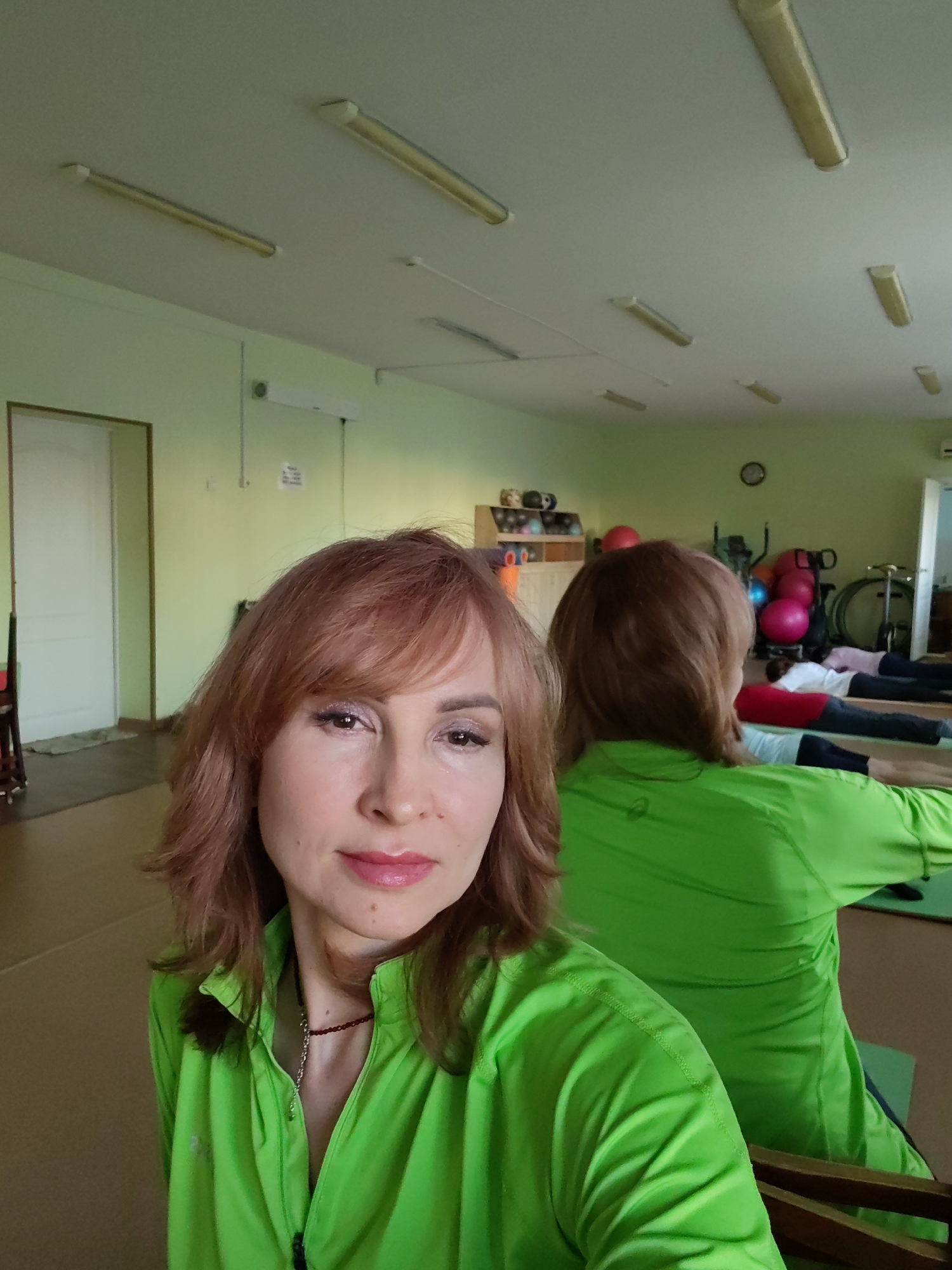 A woman with reddish-brown shoulder-length hair wearing a bright green zip-up shirt is taking a selfie in what appears to be a studio or fitness room in Chaikovsky, Russia. The room has light green walls and a white ceiling with fluorescent tube lights. In the background, several people are lying on mats, seemingly participating in an exercise class. Exercise balls, a bicycle, and other fitness equipment are visible on a shelf in the background. A white door is also present in the background. The woman is facing the camera with a neutral expression. A necklace is visible beneath the shirt.