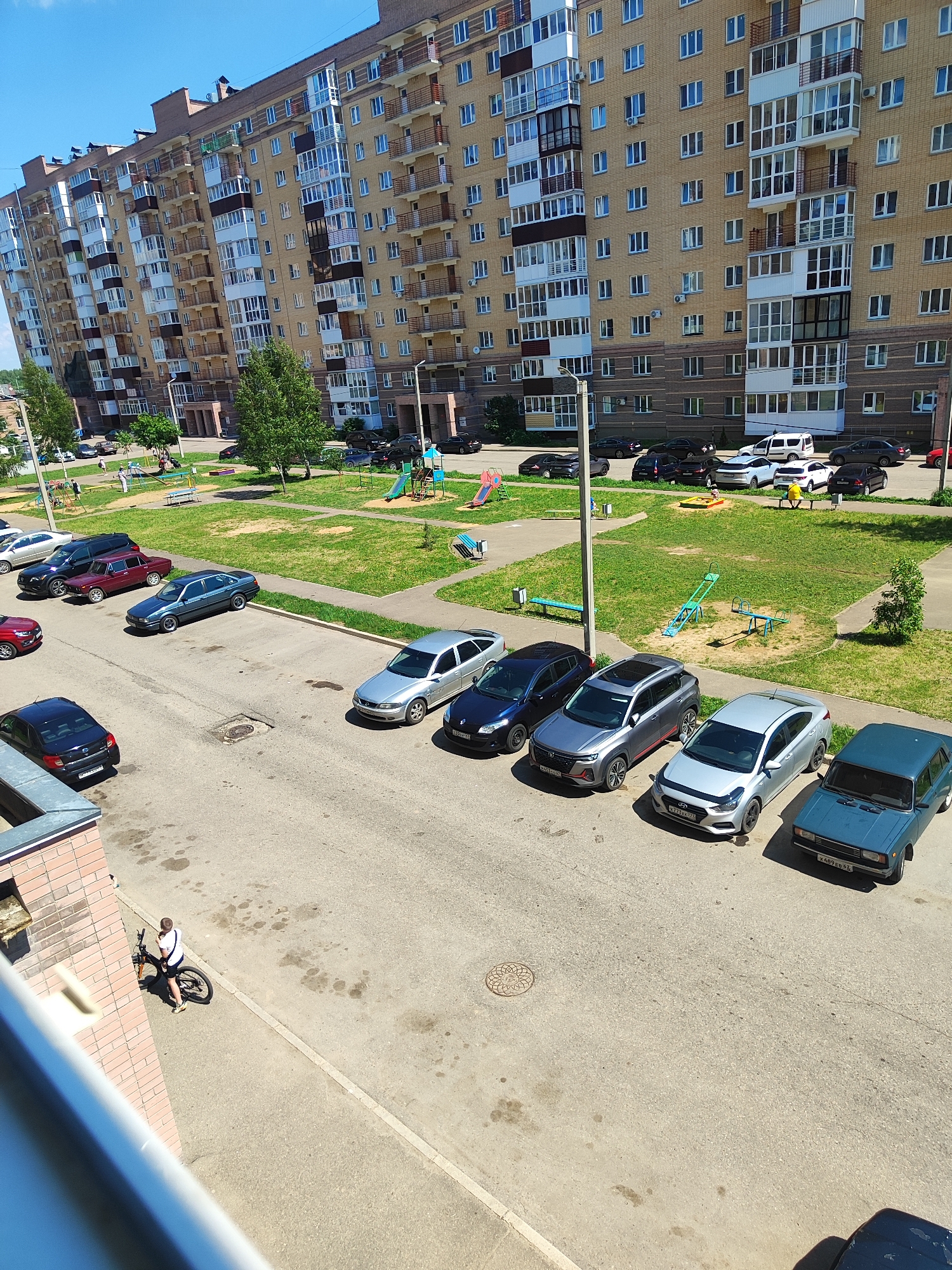 A wide shot shows a residential area with a large apartment building and a parking lot. The apartment building is tall and made of yellow brick with numerous balconies and windows. In front of the building is a green grassy area with playground equipment including swings and a slide. Adjacent to the grassy area is a paved parking lot with several cars parked. A person on a bicycle stands near a brick building in the foreground. The sky is clear and blue.