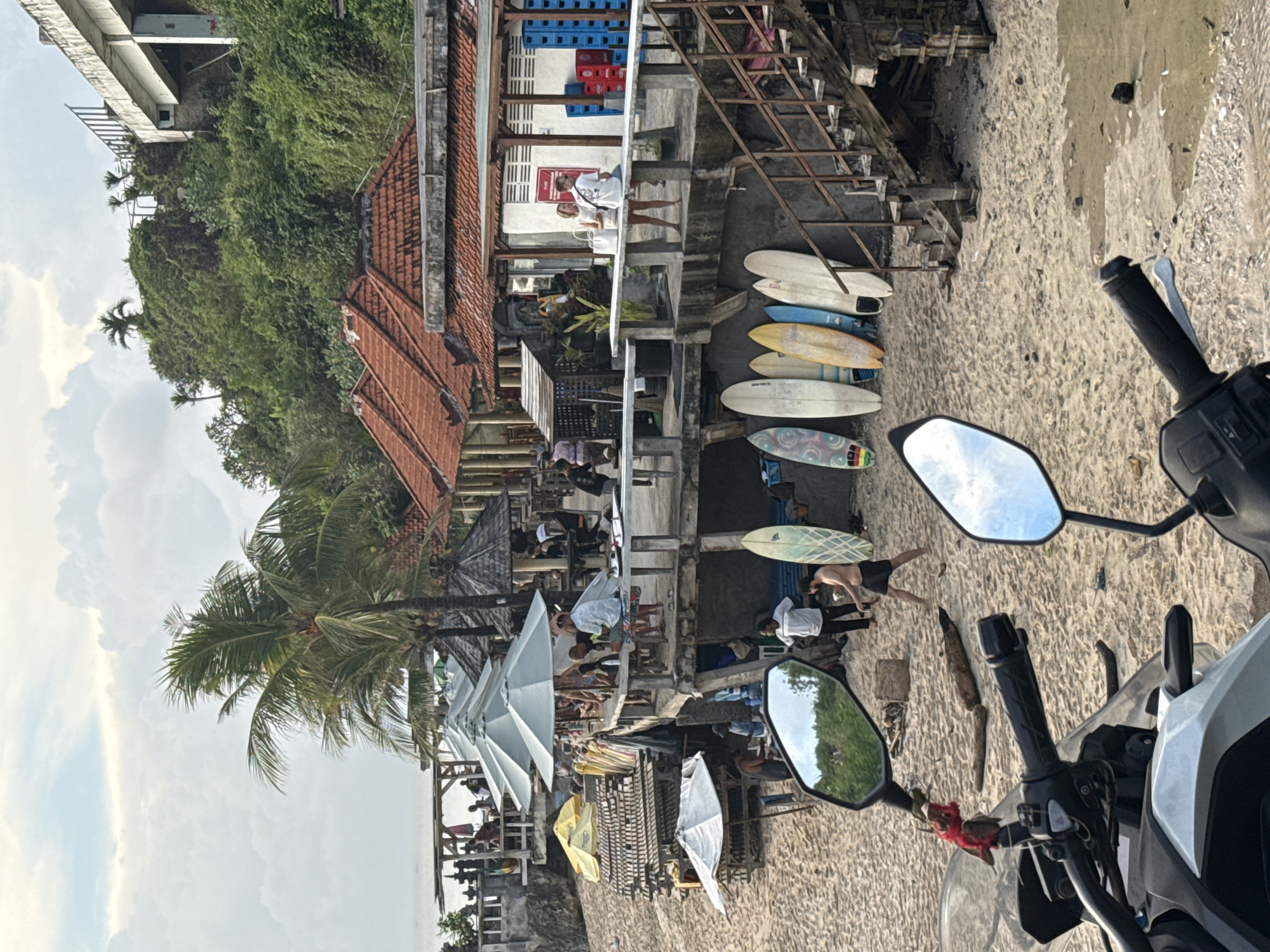 The image depicts a beach scene in Kuta Selatan, Indonesia, viewed from the perspective of someone on a motorcycle. The foreground features the motorcycle's handlebars and mirrors, reflecting the sky and surrounding trees. Beyond the motorcycle, the sandy beach is visible, with several people sunbathing, walking, and relaxing. Surfboards are stacked against a wooden structure. Further inland, a series of tiered buildings and structures line the beach, including open-air restaurants and shops with thatched roofs, umbrellas, and people dining. Dense green vegetation covers the hillside in the background. The overall impression is of a busy, tropical beachfront with a mix of recreational and commercial activity.