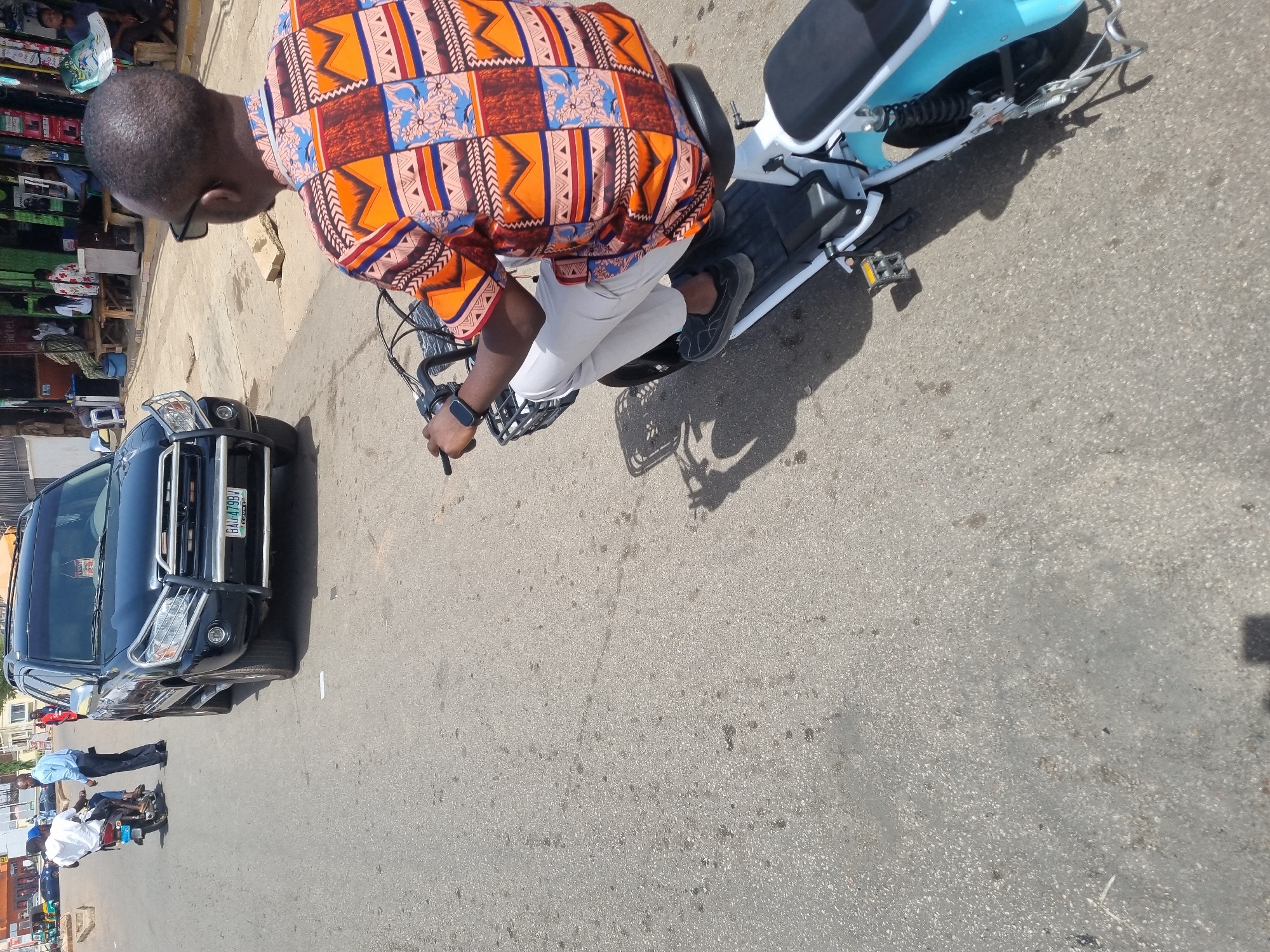 The image shows a street scene in Jos, Nigeria. A man wearing glasses, a colorful patterned shirt, and light-colored pants rides a light blue electric scooter. He is on the right side of the frame, moving from left to right. To his left, a black SUV with the license plate "BAU 479BV" is parked. In the background, on the left, there's a storefront with various stacked products. Further down the street, near the SUV, a few pedestrians are visible. The road is paved and has some discoloration. The photo is taken from an elevated perspective, looking down at the scene.