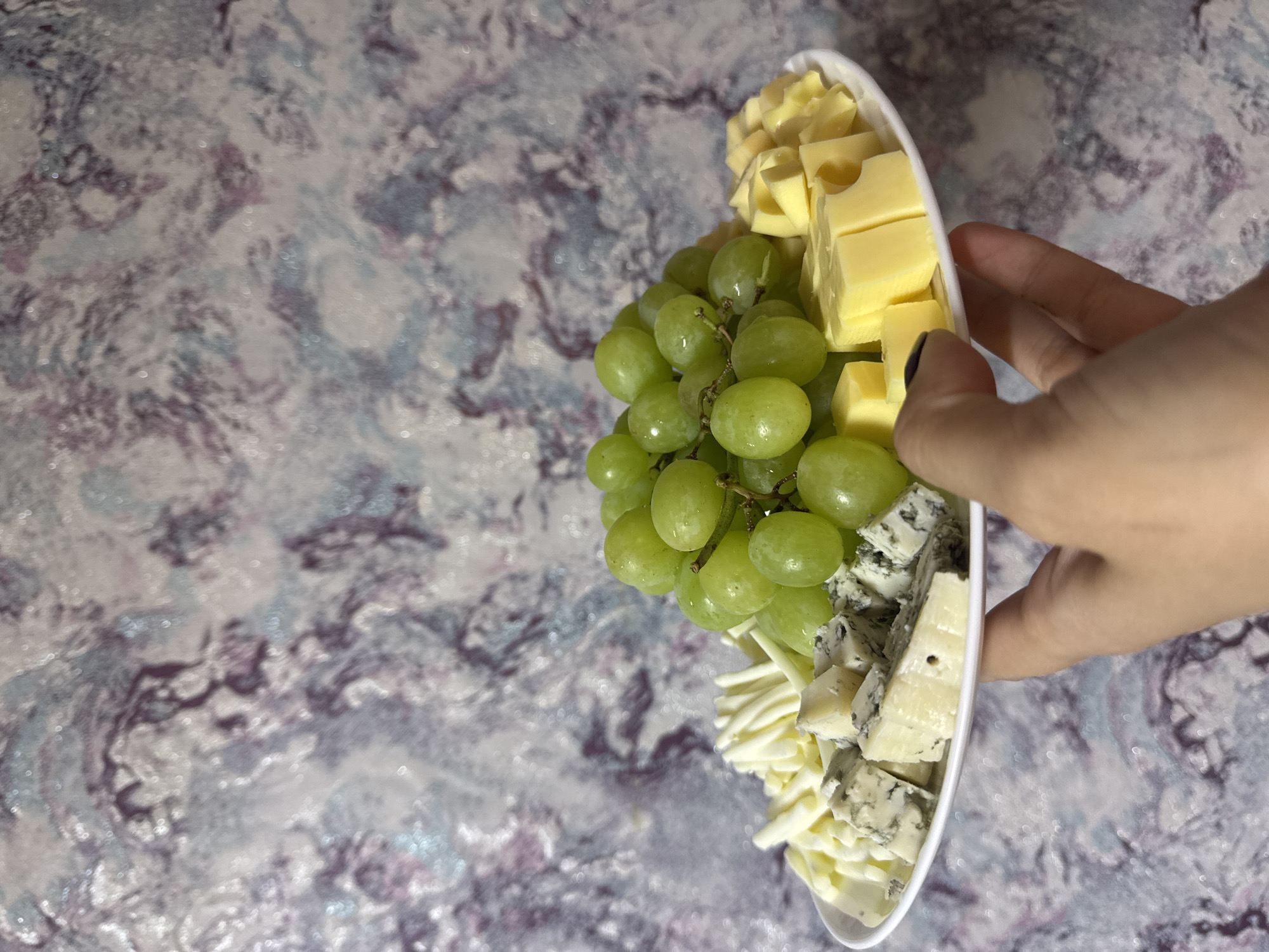 A white plate of cheese and green grapes is held by a person's hand. The plate contains cubed yellow cheese, bunches of green grapes, shredded white cheese, and cubed blue cheese. The hand is a fair-skinned hand with dark nail polish on one finger. The plate is positioned on a marbled surface with colors of purple, gray, and white. The lighting suggests an indoor setting, possibly daytime. The image is a close-up shot, focusing on the food and the hand. No text is visible in the image.