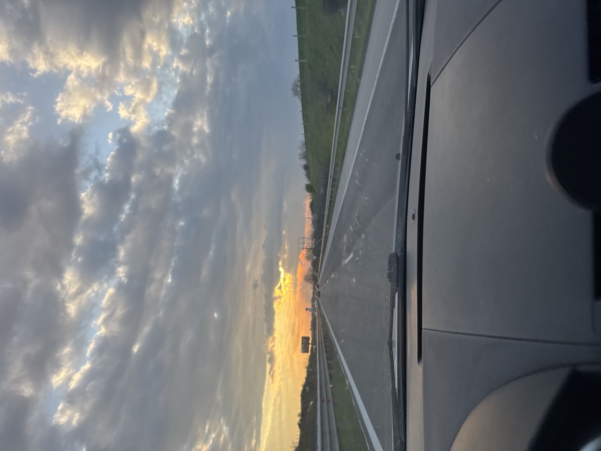 The image depicts a view from inside a car driving on a highway. The right side of the frame shows the dashboard and the edge of the car's windshield. The highway stretches into the distance with road markings visible. Guardrails are present along the side of the road. In the background, a sunset illuminates the sky with orange and yellow hues filtering through the clouds. Vegetation can be seen lining the side of the highway. No people are visible. Given the specified location of Orizovo, Bulgaria, the road is likely a highway within that region.