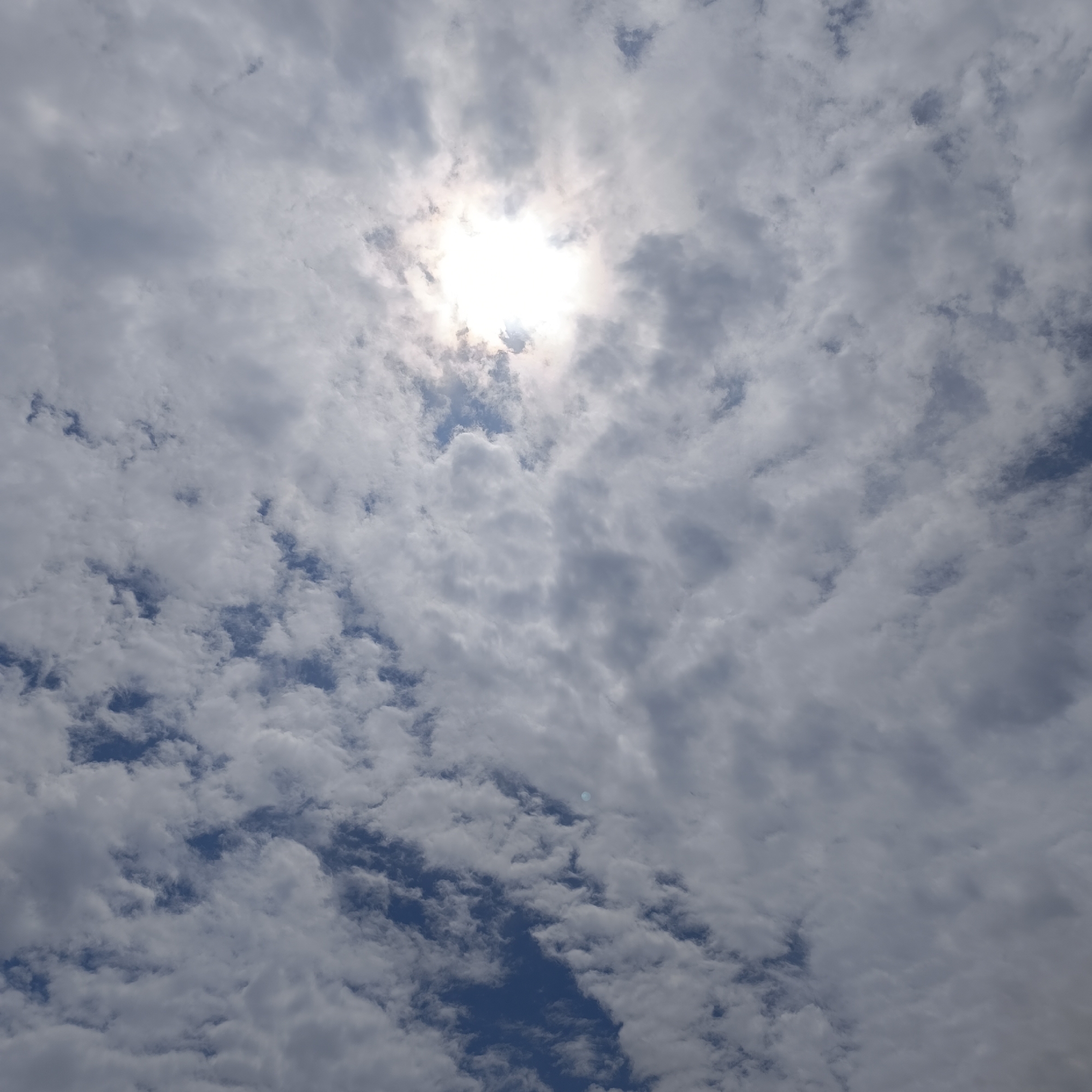 The image captures a sky filled with scattered clouds, ranging from bright white to darker gray. The sun is partially obscured by the clouds, creating a bright, hazy halo effect at the top center of the frame. Patches of blue sky are visible between the clouds, suggesting it is daytime. The overall impression is of a partly cloudy day, likely with a moderate amount of sunlight filtering through. There are no people, objects, or discernible location cues other than the sky itself. No text is visible in the image.