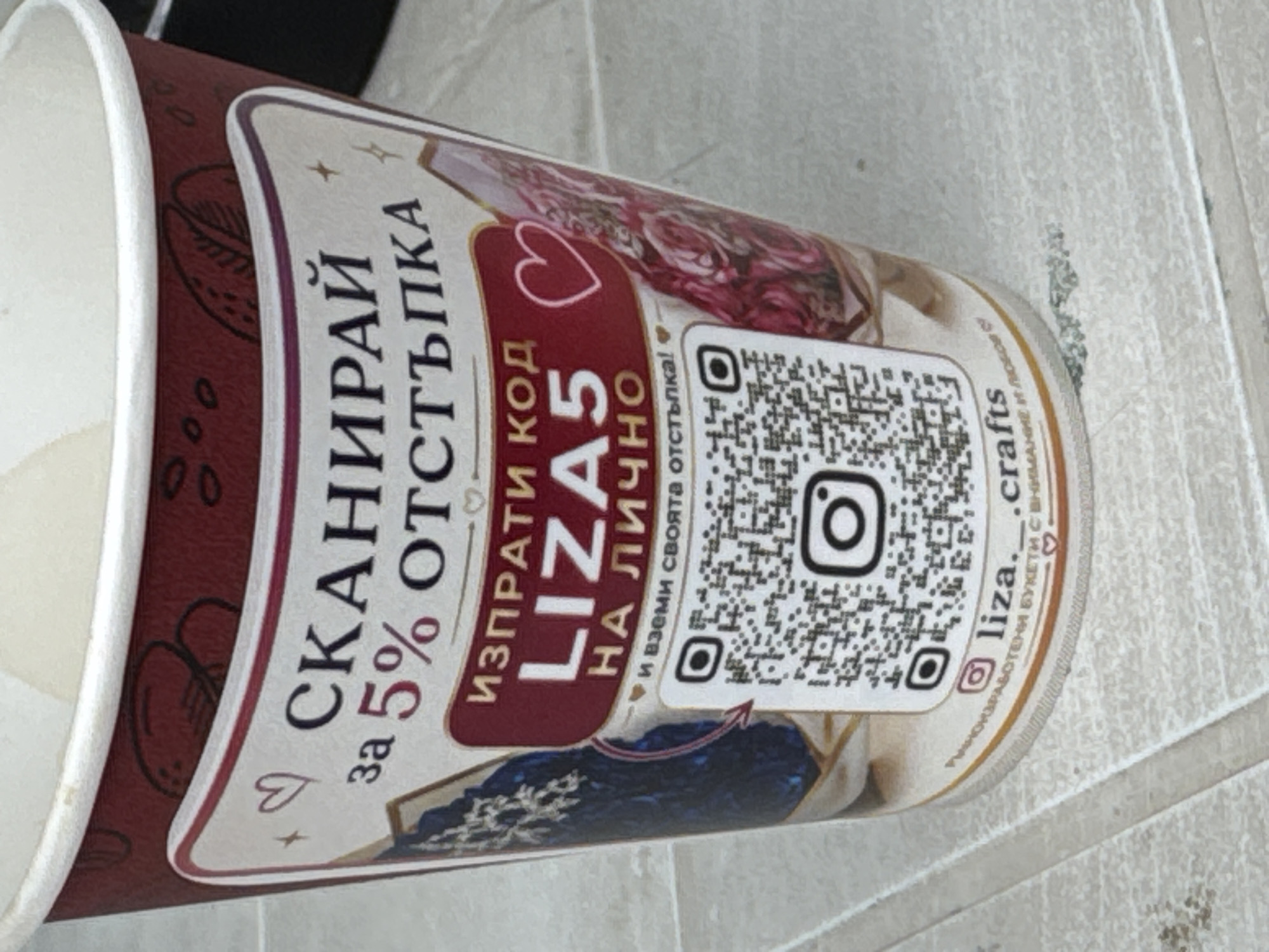 The image shows a close-up of a paper cup with a promotional design. The cup is primarily dark red with a cream-colored rim. The design features text and graphics, including a QR code and social media icons. The text, in Cyrillic script, appears to be an advertisement for a discount. Specifically, it reads "СКАНИРАЙ за 5% ОТСТЪПКА" which translates to "SCAN FOR 5% DISCOUNT". Below this, it says "ИЗПРАТИ КОД LIZA5 НА ЛИЧНО" meaning "SEND CODE LIZA5 PRIVATELY". There are also smaller text elements, including an Instagram logo and the handle "liza.__crafts", and a phrase "РЪЧНОРАБОТЕНИ БУКЕТИ С ВНИМАНИЕ И ЛЮБОВ" which translates to "HANDMADE BOUQUETS WITH CARE AND LOVE". The background is a light-colored, textured surface that appears to be a table or countertop. There are no people or animals visible in the image. The location is not identifiable from the provided image.