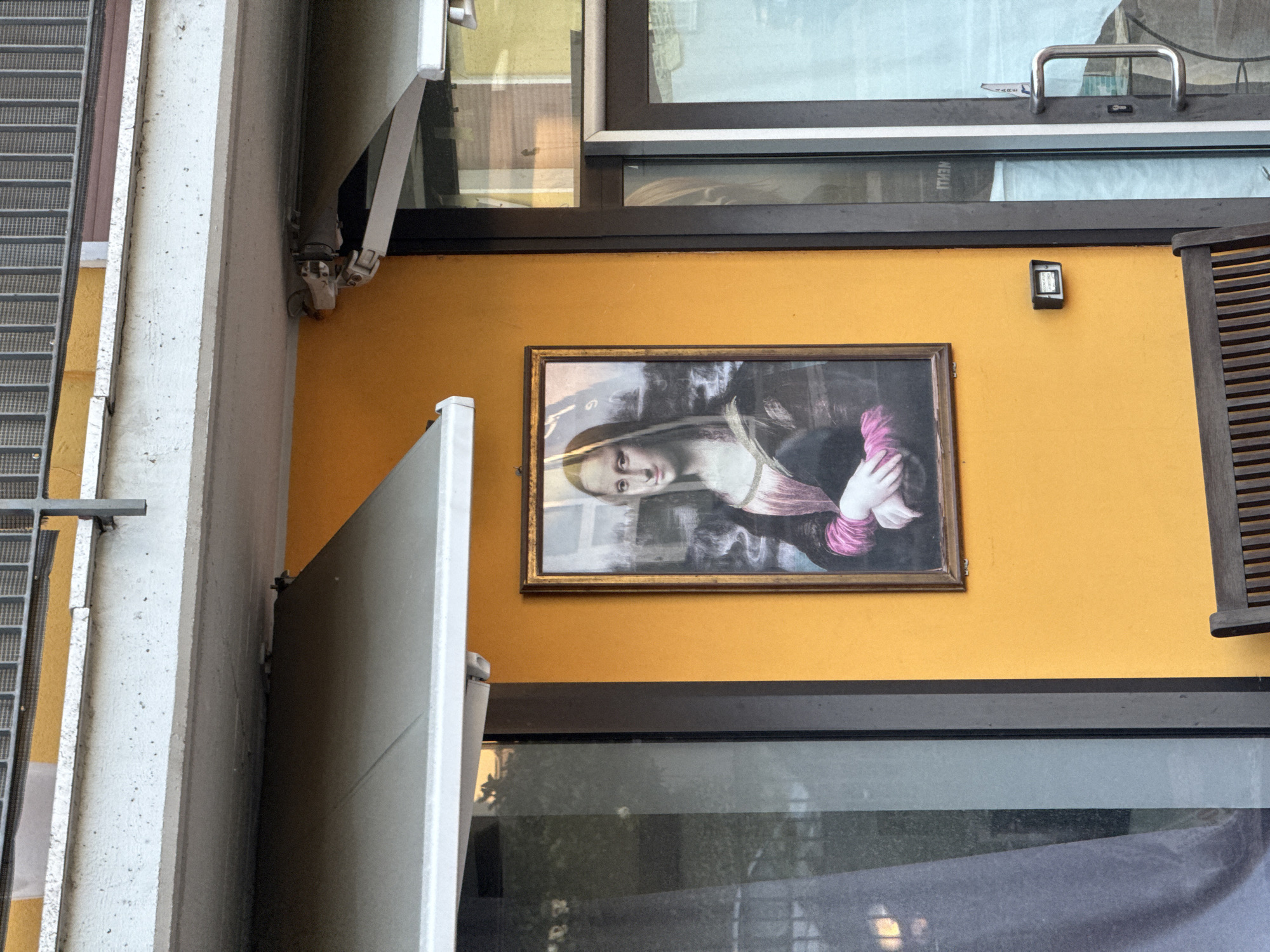 A building exterior is visible. The facade is yellow, and a framed painting of a woman is mounted on the wall. A closed white door is beside the painting, and a window is above. A security device is also on the yellow wall. The location is Villafranca di Verona, Italy.