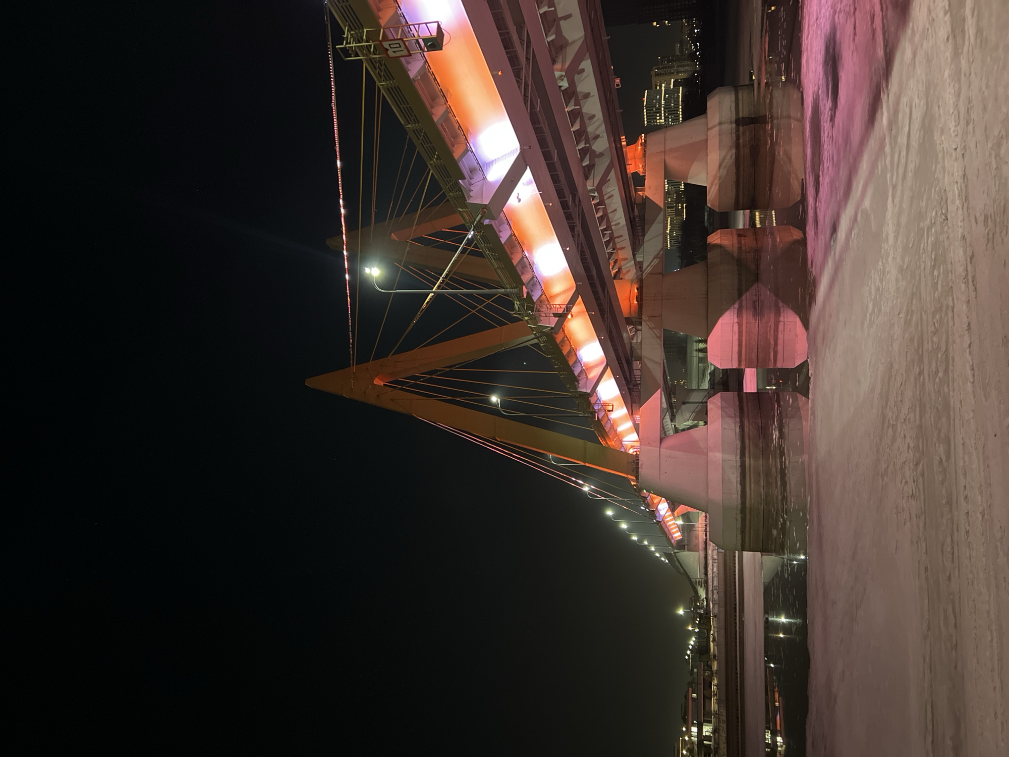 The image depicts the Millennium Bridge in Kazan, Russia, at night during winter. The cable-stayed bridge features a prominent yellow-orange M-shaped pylon visible on the right, supporting the bridge deck with numerous cables. The bridge's underside is illuminated with alternating orange and purple/pink lights. Streetlights line the bridge deck, and red string lights are visible along the upper structure. A sign displaying the number "10" is affixed to the bridge infrastructure. Below the bridge, the Kazanka River is partially frozen, displaying expanses of snow and ice alongside areas of open water. The bridge's illumination reflects off both the water and the snow-covered ice, casting pink and purple hues onto the foreground. Concrete bridge support columns are visible emerging from the water. In the distant background, illuminated city buildings mark the opposing riverbank under a dark, clear sky. No individuals or vehicles are present in the scene.