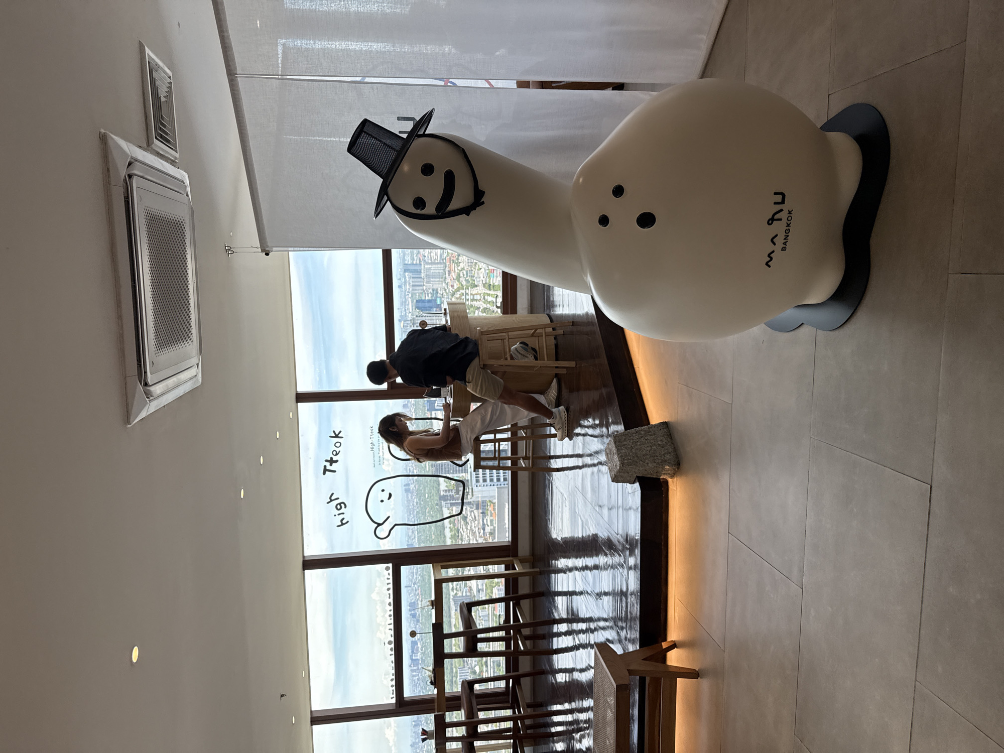 The image shows a high-angle interior shot with a large, whimsical sculpture dominating the foreground. The sculpture consists of two white, rounded forms, one shaped like a bowling pin with a top hat and drawn-on face, and the other a larger sphere with three dots arranged like eyes, and the brand name "Manu Bangkok" printed on its side.
In the background, floor-to-ceiling windows reveal a cityscape, implying a high elevation within a building. Two people are partially visible in front of the windows, one wearing a dark shirt and the other a light top. They appear to be interacting, possibly engaging in a transaction or discussion at a counter or table.
Scattered chairs and tables suggest a cafe or lounge setting. "high Tteok" is written in a childlike, cursive font on one of the window panes. Light fixtures are visible on the ceiling.
