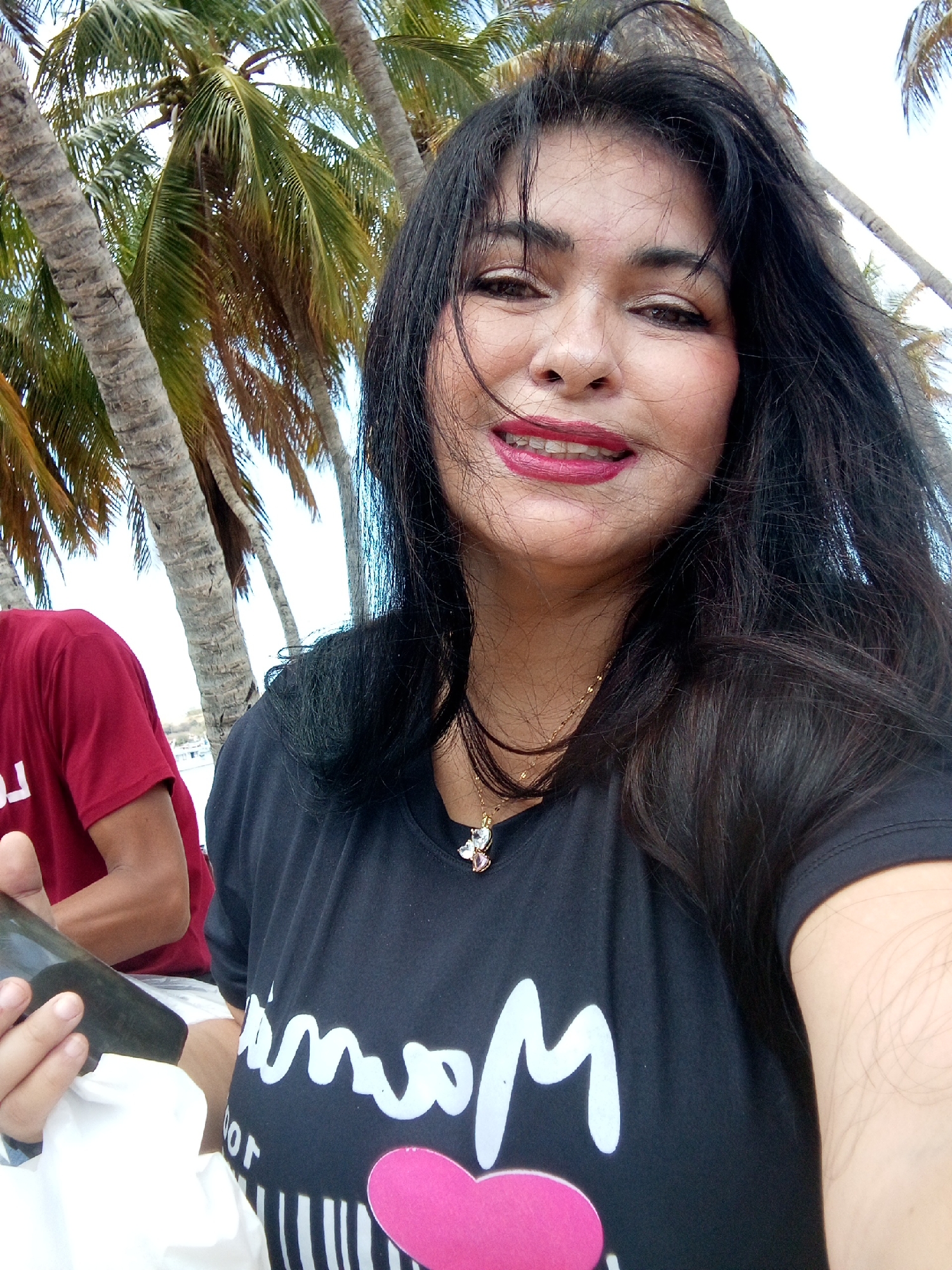 A medium shot captures a woman in the foreground, with palm trees suggesting a tropical beach location. The woman is wearing a dark t-shirt with white writing and a pink heart. She is also wearing a necklace. She appears to be holding a dark object. To the left of the frame, a person in a red shirt is partially visible. The background includes more palm trees and the sky. The location seems to be a beach, given the palm trees and sky. The city is Unknown, Unknown.