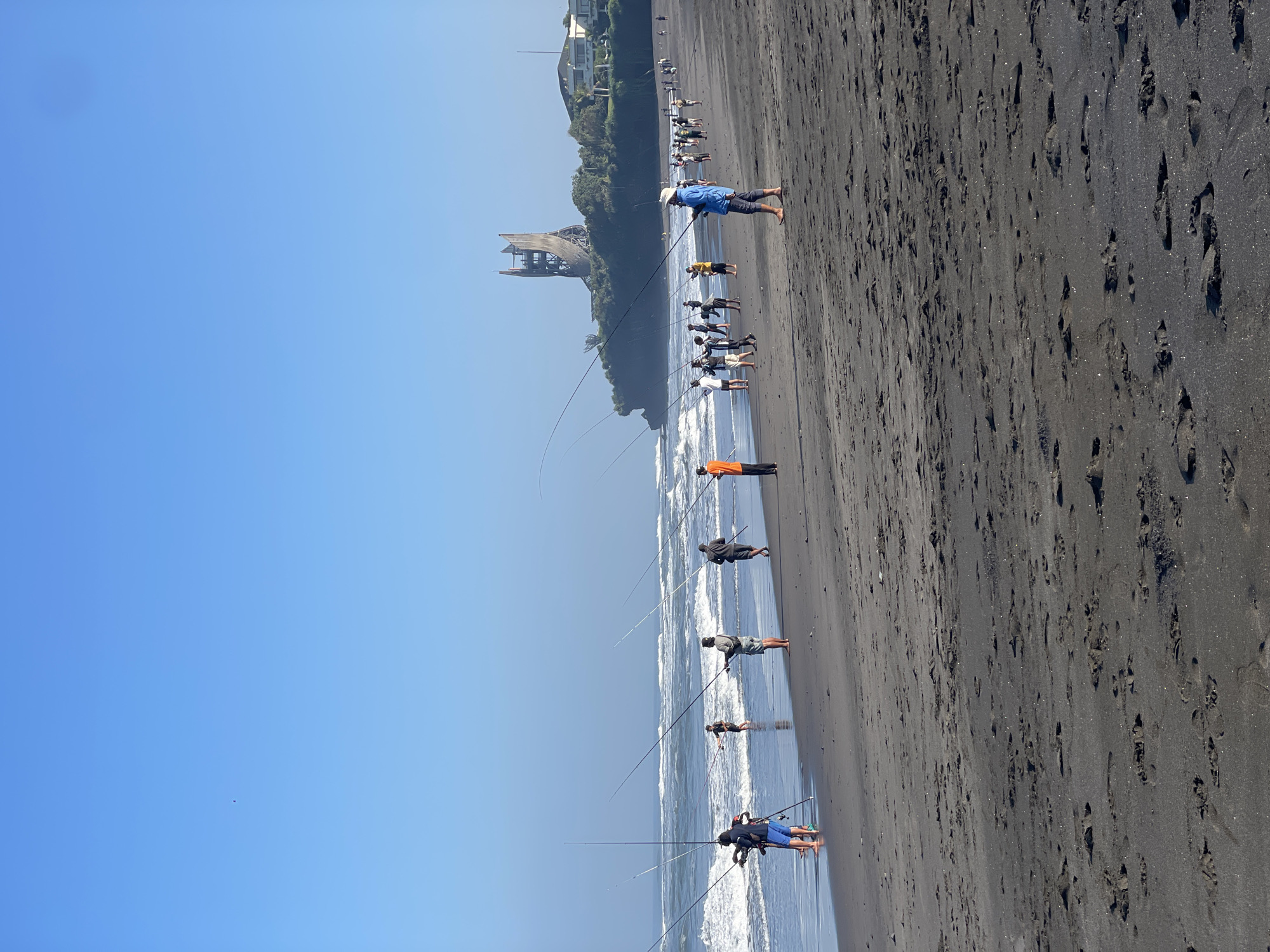 The image depicts a wide, sandy beach with dark sand, characteristic of some Indonesian coastlines. The ocean is active with moderate waves breaking on the shore. A group of people, estimated to be around 20-30 individuals, are spread along the beach, actively engaged in fishing with long fishing rods. They are positioned at varying distances from the water's edge, with some standing in the shallow surf and others on the drier sand. The individuals are dressed in casual beachwear.
In the background, a distinctive, partially constructed tower-like structure is visible on a rocky outcrop covered in vegetation. Adjacent to this, a modern building complex with white walls and a tiled roof can be seen. The sky is clear and blue, indicating fair weather. The scene conveys a sense of community activity focused on recreational fishing on a coastal day. The specific location in the city of Kediri, Indonesia, is not directly evident from the visual elements alone; however, the presence of black sand and the architectural style of the structures in the background are common features found along the southern coast of East Java, where Kediri is located. This image captures a moment of shared leisure and outdoor pursuit.