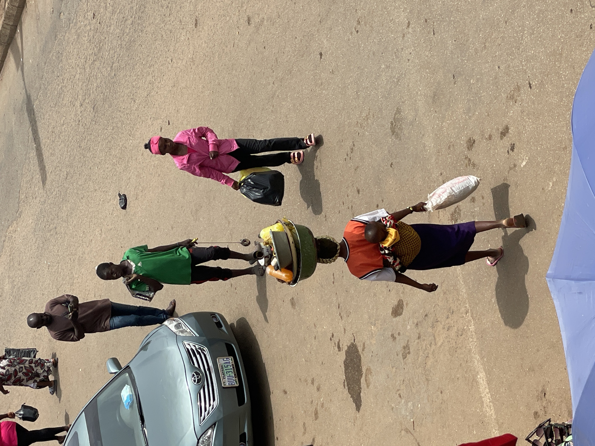 The image shows a street scene in Bukuru, Nigeria. A light blue Toyota car with license plate "BUU-315AG" is parked on the side of the road. Several people are present. A woman in a pink jacket and black pants is carrying a black plastic bag. A man in a brown shirt and blue jeans is looking at his mobile phone. A man in a green shirt is also looking at his phone. A woman is carrying a large tray of goods on her head, while also carrying a small child in her arms and what appears to be a small bag in her hand. A blue umbrella is visible at the right. There are also several other people present, but they are obscured. The scene appears to be taken from an elevated perspective.