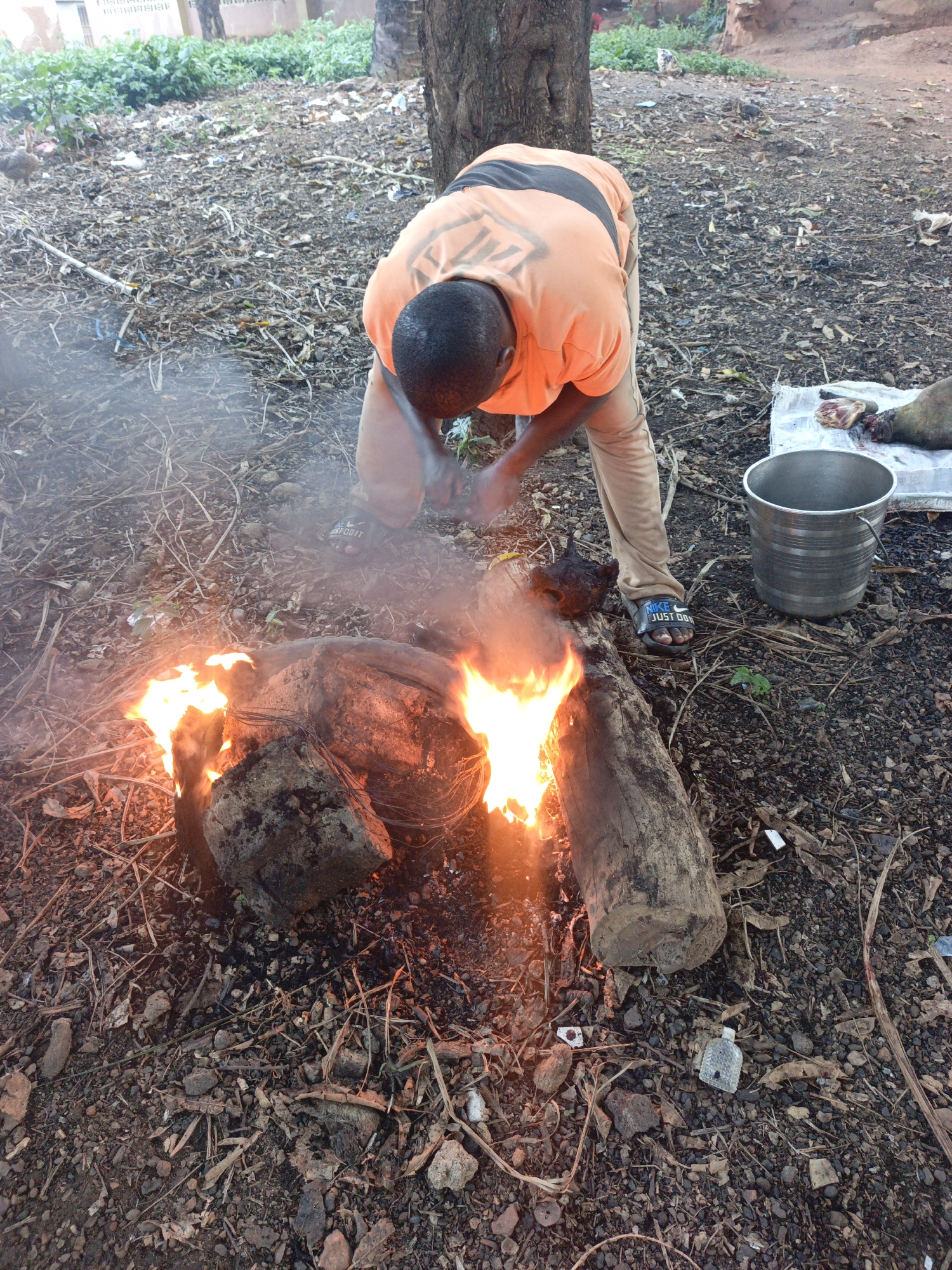 In Akwanga, Nigeria, a man wearing an orange shirt and tan pants is bent over a fire, holding an object. The fire is burning wood. Next to the man is a silver bucket. A white cloth is on the ground with what appears to be animal parts on it. The ground is littered with leaves, twigs, and rocks. In the background, there is some greenery and a building.