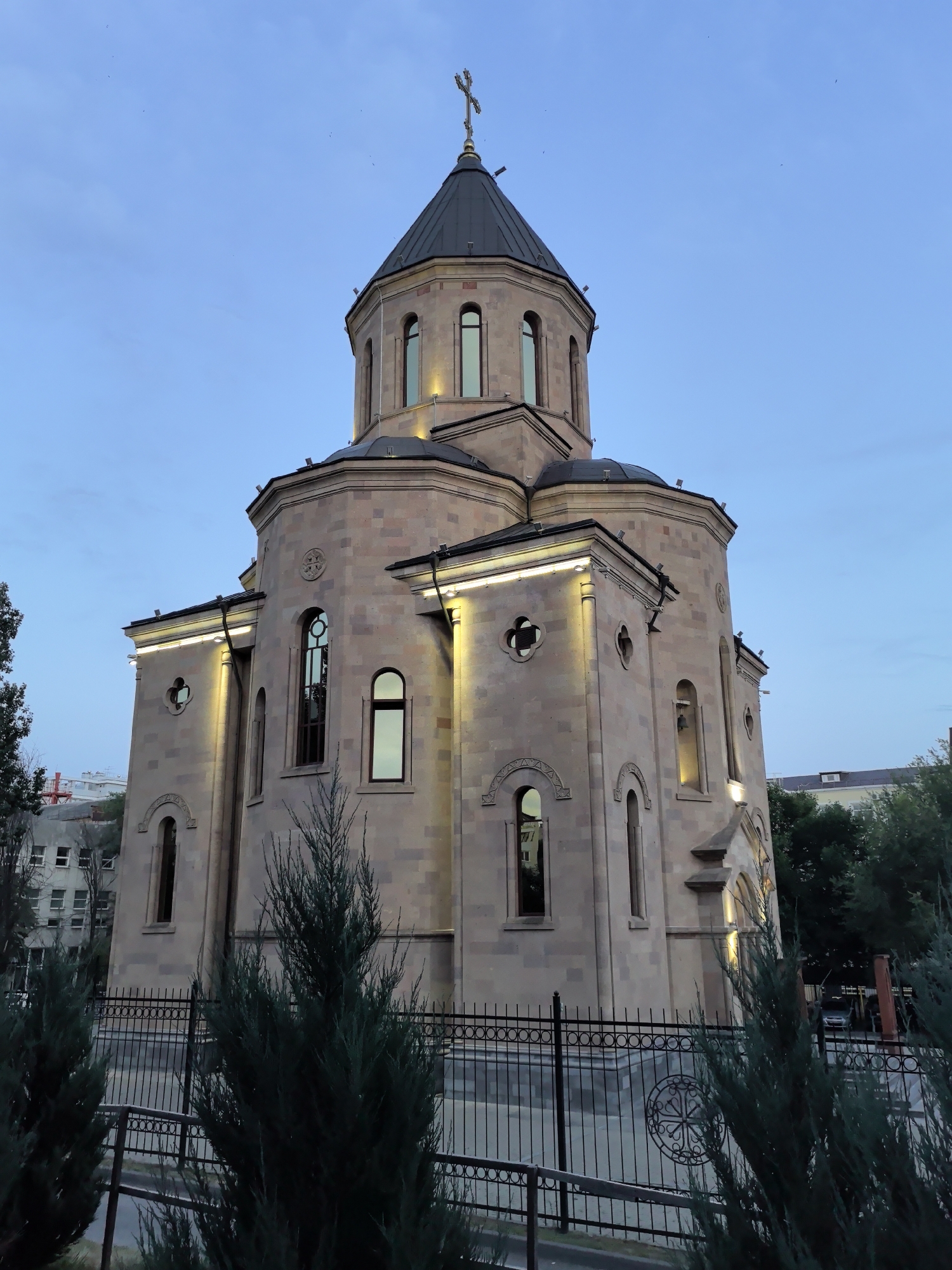 An external, low-angle, full-shot photograph captures a stone church with a dark, conical dome topped by a cross. The church's facade is illuminated by several exterior lights. The sky is clear and a pale blue, indicating either dawn or dusk. The church is surrounded by a black metal fence, in front of which are several evergreen trees. In the background, the upper floors of a multi-story building are visible, with trees and other structures interspersed. The image appears to be taken in the city of Veshenskaya, Russia.