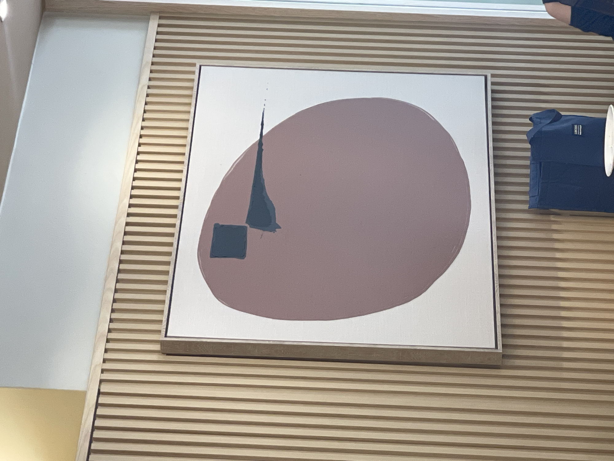 The image shows a framed abstract painting on a wall paneled with vertical wooden slats. The painting features a large, muted pink oval shape with a dark blue square and a dark blue stroke across it. Below the painting, a dark blue tote bag is visible. The environment appears to be an indoor setting, possibly a cafe or gallery, with a light-colored wall and ceiling. The exact location in Kuala Lumpur, Malaysia cannot be determined from the image.