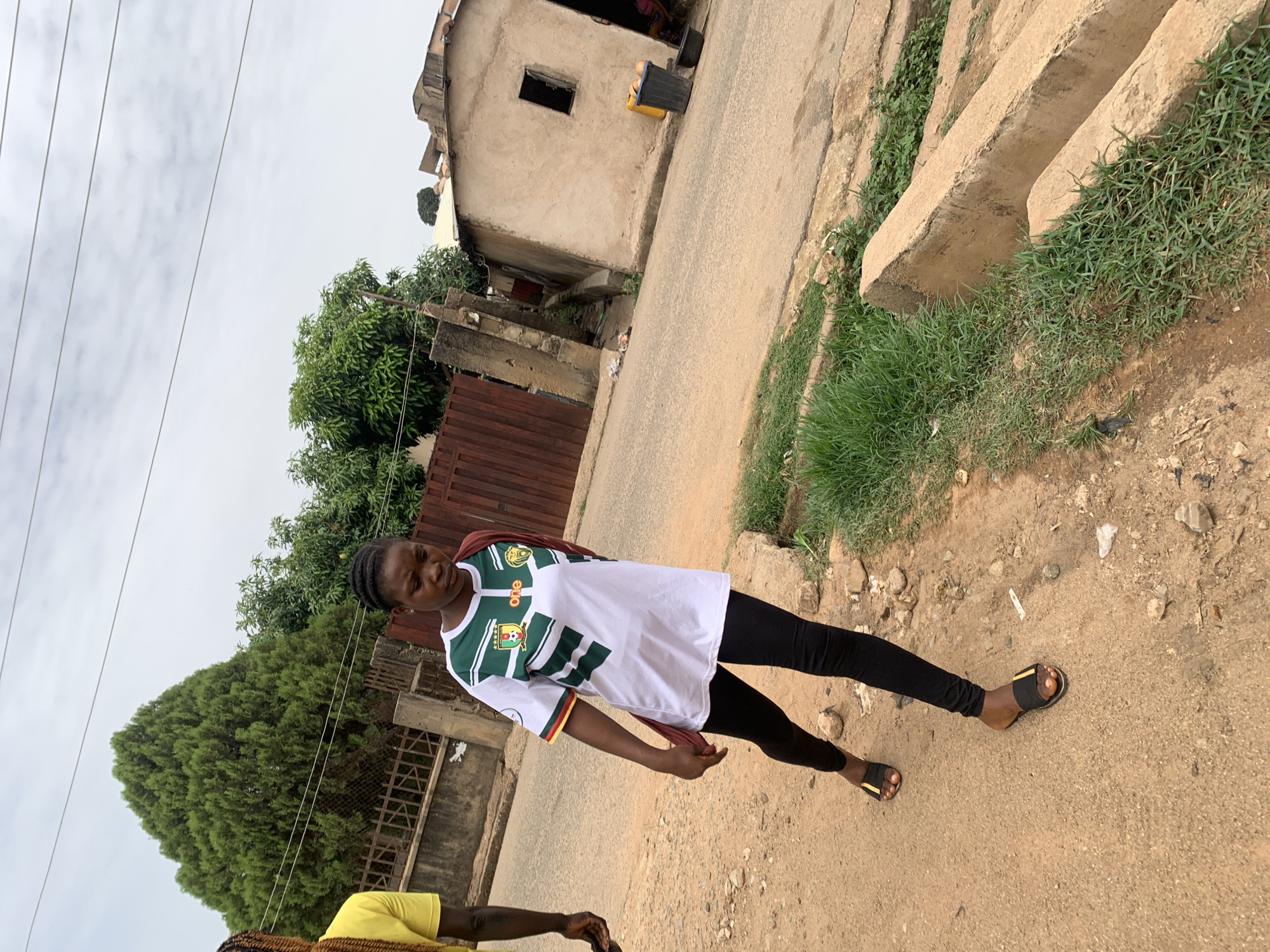 A young woman stands in the street in Gura Top, Nigeria. She wears a white t-shirt with green and yellow accents and black leggings. The street is unpaved, with vegetation on the sides. A simple, one-story building is visible in the background. The sky is overcast.