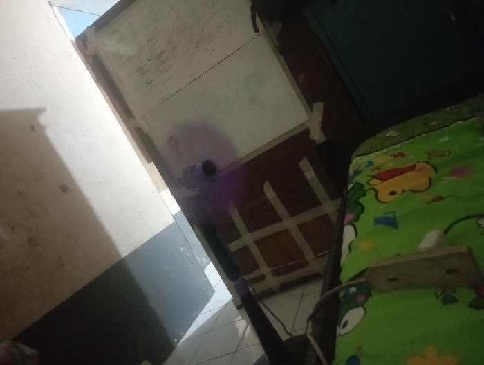 A cluttered room shows a partially visible whiteboard with writing on it. A fan stands nearby, casting a purple glow. A bedspread featuring Winnie the Pooh characters is prominent in the lower right corner, with a power strip resting on top. The room is dimly lit, and the angle is slightly skewed, adding to the disorganized feeling.