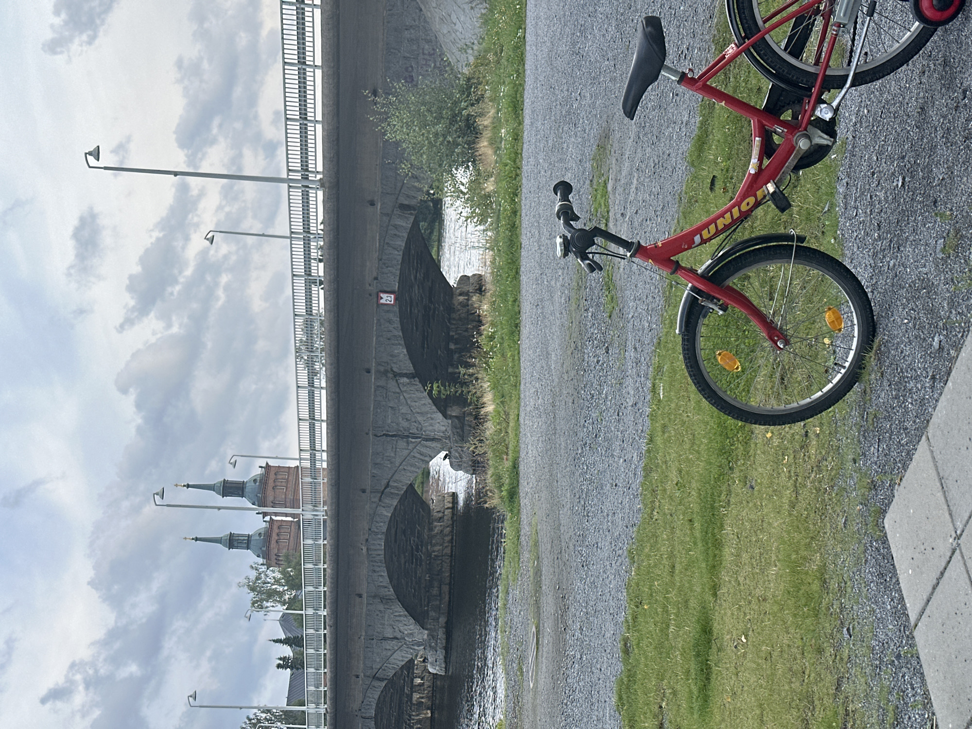 The image depicts a red child's bicycle parked on a gravel path next to a grassy area. In the background, a stone bridge with multiple arches spans a body of water. Beyond the bridge, two church towers with green spires are visible against a cloudy sky. The overall scene appears to be an outdoor public space in Vammala, Finland, likely near a river or canal.