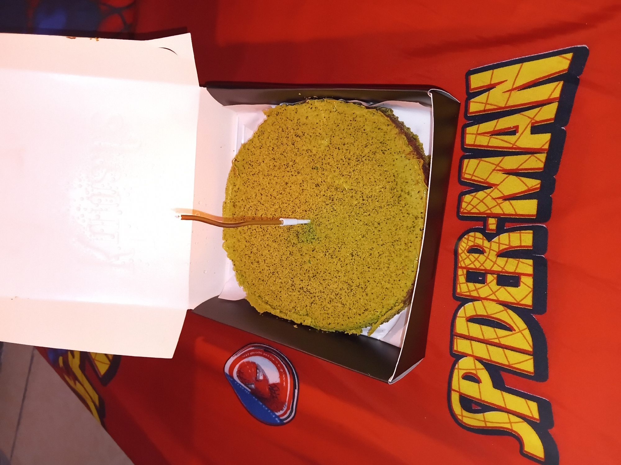 A round, green-frosted cake with a single candle is centered in a black cardboard cake box, which is open. The cake box is placed on a red tablecloth that features a large, yellow "SPIDER-MAN" logo in the lower half. To the left of the cake, a circular patch with a Spider-Man emblem is also visible on the tablecloth. The background is out of focus. This scene is likely from a birthday celebration, given the cake and candle.