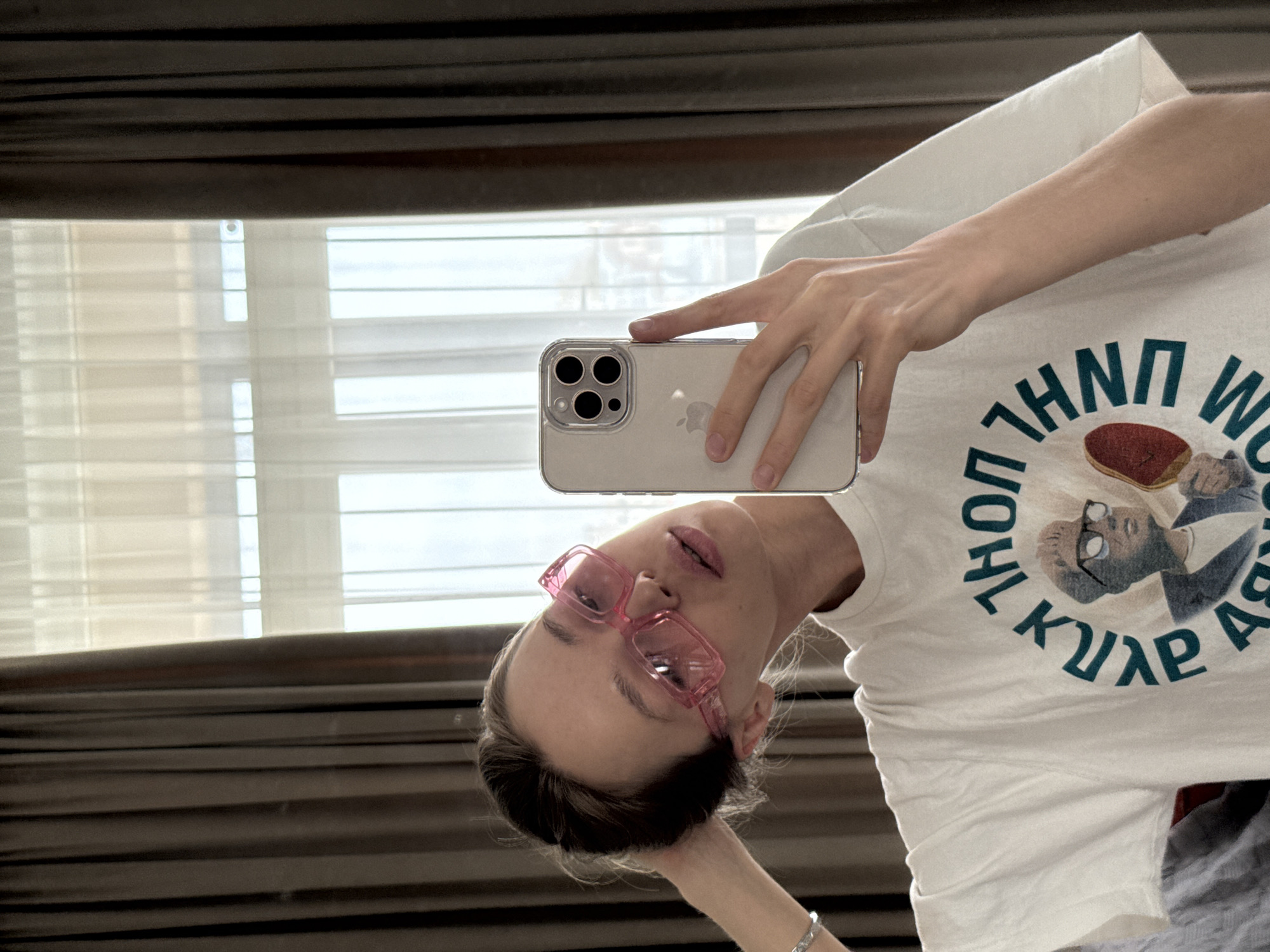 A person is captured in a mirror selfie, with the image rotated 90 degrees counter-clockwise. The subject, likely female, has dark hair pulled back and is wearing distinctive pink, rectangular-framed glasses. She looks towards the camera with a neutral expression.
She holds a light-colored smartphone, which appears to be an iPhone with a triple-lens camera system, enclosed in a clear case. Her left hand rests near her head, showcasing a silver-colored bracelet on her wrist. She is wearing a white t-shirt featuring a prominent circular graphic design on the chest.
The graphic on the t-shirt displays a cartoonish figure of an older man with glasses and gray hair, dressed in a suit, holding a red, heart-shaped or ball-like object. Surrounding this figure is stylized, reversed text in a green color, arranged in a circular fashion. The visible letters, when un-reversed, suggest words or names, possibly in a non-Latin script, such as "WORLD" or "HUNTER" on the upper part, and "АРЯЦК" (ARYACK) on the lower part, but are illegible in the mirrored state.
The background consists of light-filtering window blinds, partially open, allowing bright daylight to enter, indicating a daytime setting indoors. The blinds appear to be horizontal and have a dark brown or black top valance.