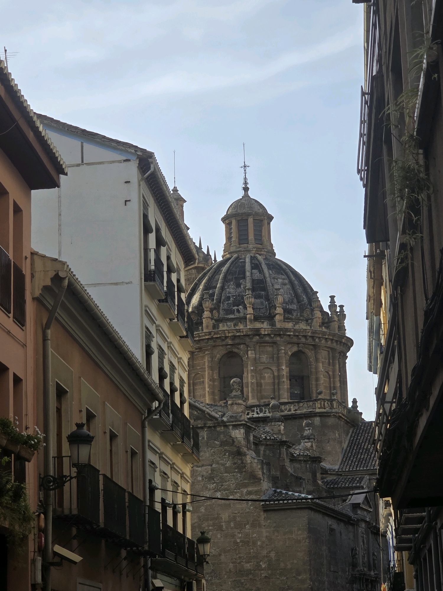 The image shows a narrow street in Granada, Spain, with an old, ornate dome of a building visible between the structures on either side. The buildings lining the street are multi-story, with balconies, windows, and awnings. One building on the left is painted orange, while the one next to it is white. The building on the right is dark and appears to have plants growing on its exterior. The sky above is a pale blue with some clouds. The dome in the background is made of stone and features intricate architectural details, including arches, decorative elements, and a cross at its apex. The scene suggests an urban environment with historic architecture. There are no people visible in the image.