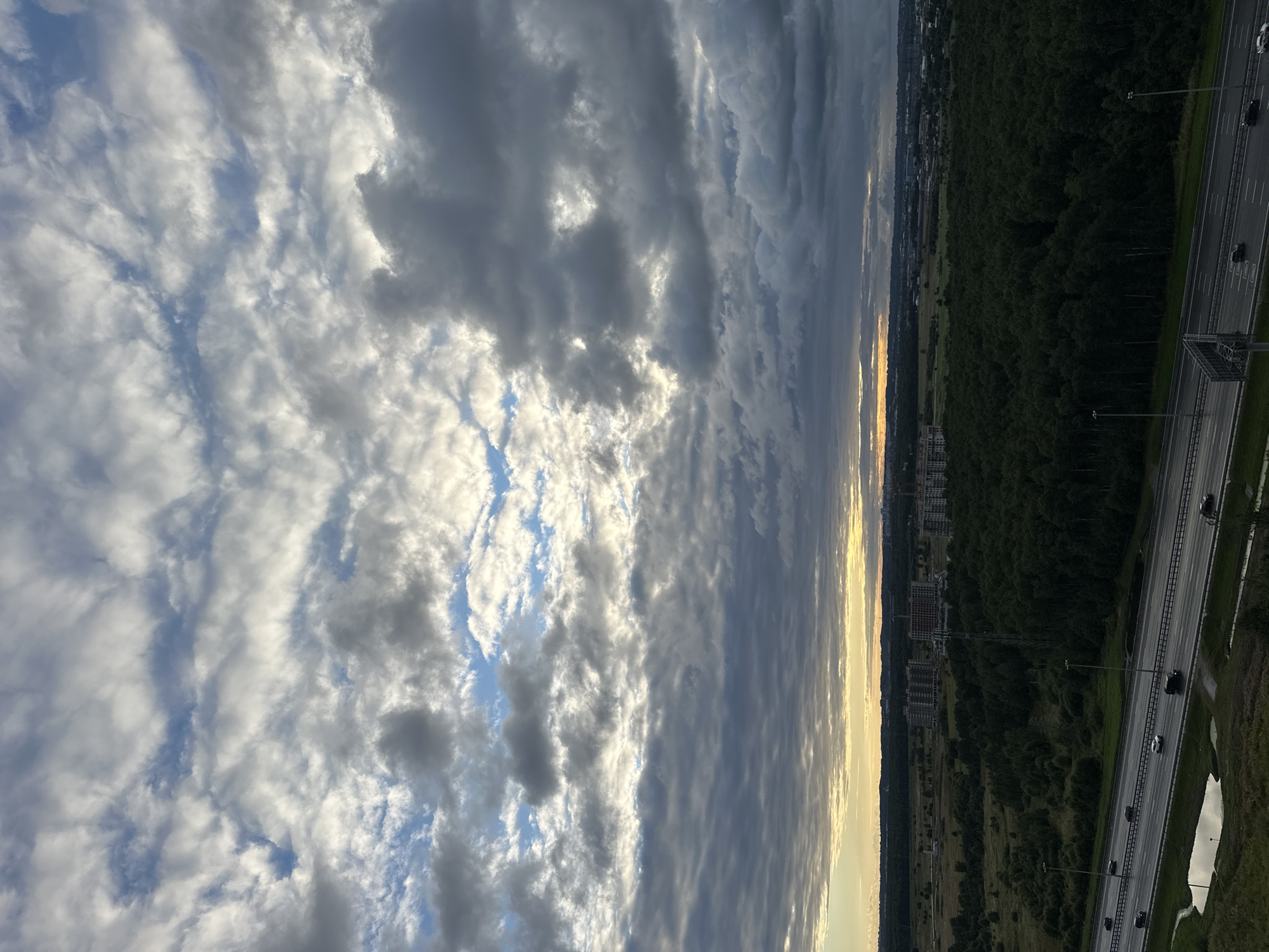 The image shows a wide, elevated view of the city of Kudrovo, Russia. The sky is filled with dramatic, textured clouds in shades of grey and white, with patches of bright blue sky visible and a muted yellow and orange glow on the horizon, suggesting either sunrise or sunset. Below the sky, a dark green, dense forest occupies the middle ground, interspersed with some grassy fields. Beyond the forest, several apartment buildings and construction sites are visible, indicating ongoing urban development. A multi-lane highway runs horizontally across the lower portion of the image, with a few cars driving on it. The road appears wet, possibly from recent rain, and a small puddle of water is present on the left side of the highway. The overall scene depicts a somewhat overcast, developing urban landscape adjacent to a large wooded area.