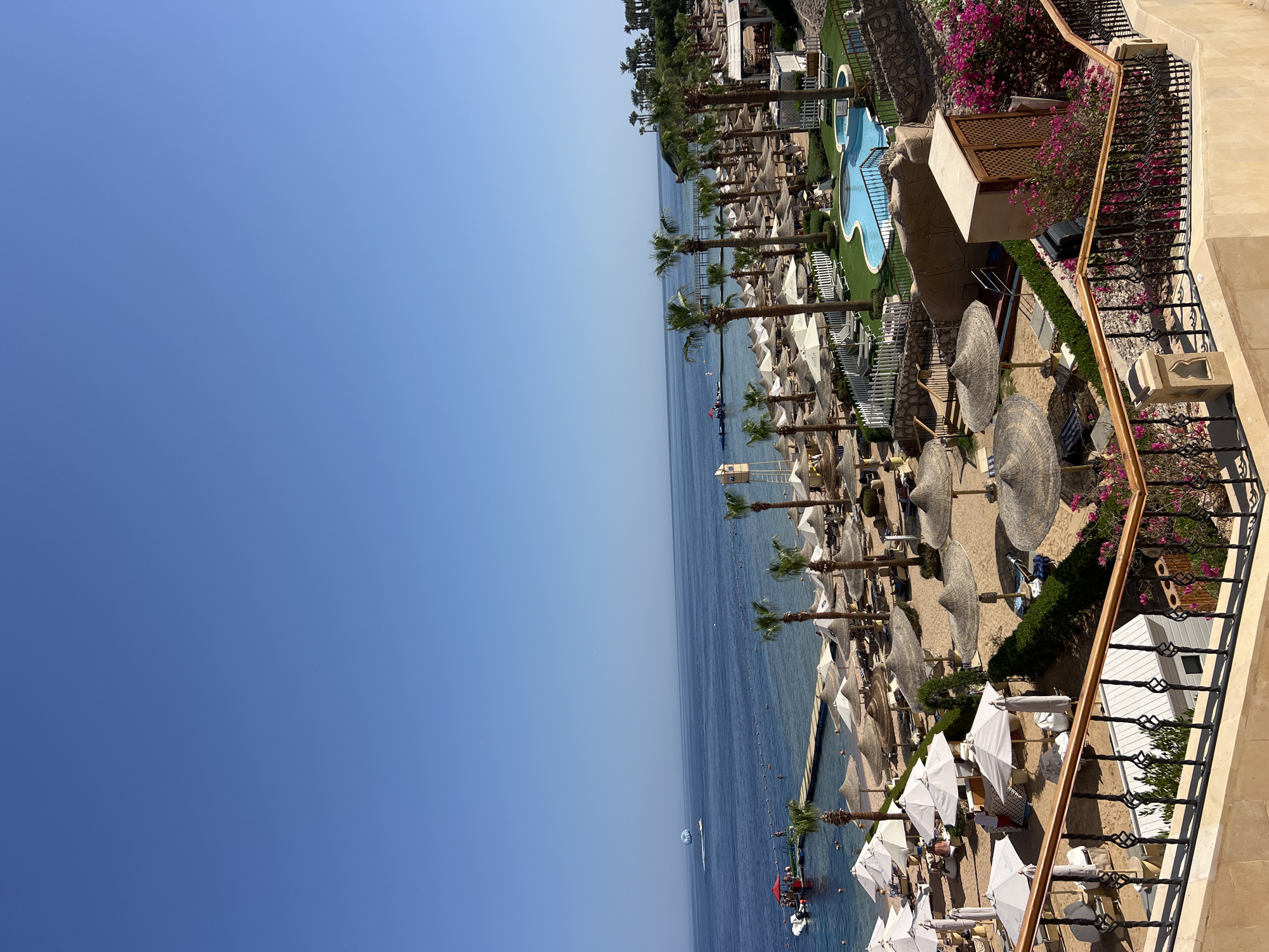 A high-angle, wide shot shows a tropical resort beach on a clear, sunny day. The foreground features a decorative balcony railing overlooking a beach lined with numerous straw umbrellas and white lounge chairs. Beyond the beach, a calm, blue ocean stretches to the horizon, with a few small boats and a parasailer visible. Palm trees are scattered throughout the resort, and a swimming pool with blue water is visible in the middle ground. The scene suggests a vacation destination in Sharm El Sheikh, Egypt.