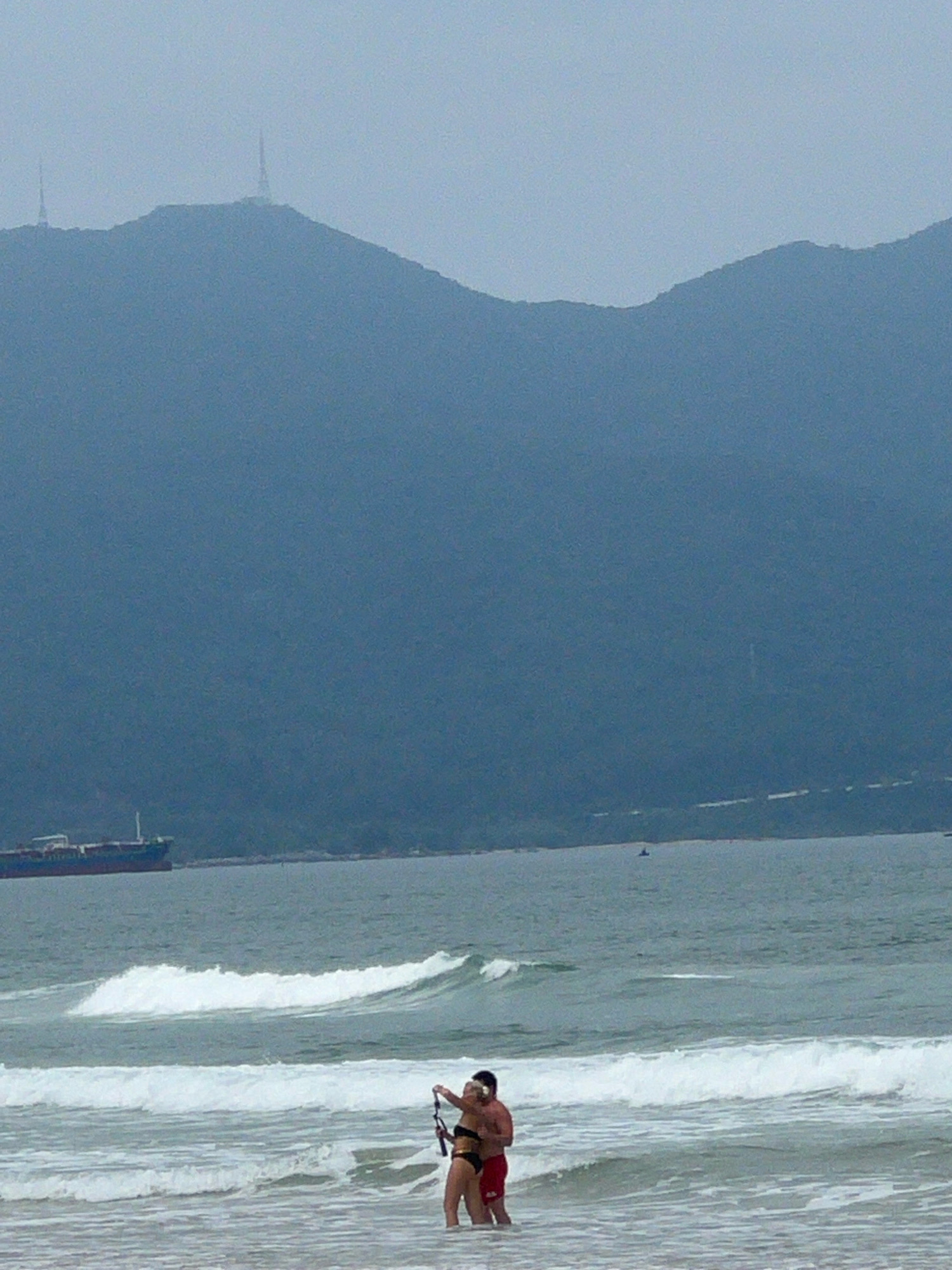 In Đà Nẵng, Vietnam, a couple stands in the shallow ocean water near a beach. The man, wearing red swim trunks, embraces the woman, who is in a black bikini. They are taking a selfie with a selfie stick. The ocean waves are breaking around them. In the background, a large cargo ship is visible on the water. Further in the distance, a lush green mountain range dominates the horizon, with two prominent telecommunications towers atop its peaks. The sky is overcast, casting a muted light over the scene.