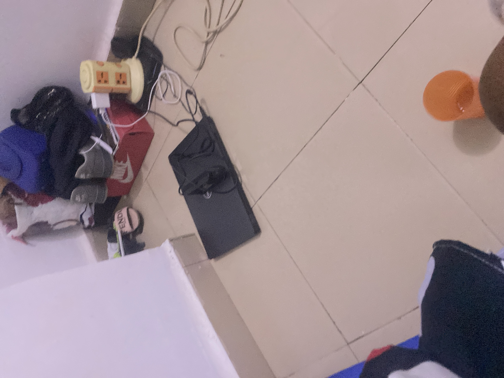 This is a photograph of a cluttered corner of a room, likely a bedroom or storage area, with a pale pink wall on the left and a tiled floor. The objects present suggest a lived-in space with items for personal use and potentially electronics.
In the background, on a shelf or elevated surface, are several items including a red shoebox with a Nike logo, a pair of grey athletic shoes, and some bags and clothing. A yellow multi-socket power adapter is plugged into the wall and is connected to various cords, including a white cord and a black cord. The black cord is connected to a black laptop which is open and lying flat on the floor in the mid-ground. The laptop has some cables plugged into it, including a black cable and a thinner cable. A pink sandal with "FENDI" written on it is visible on the floor near the shelf.
In the foreground, the tiled floor is visible, with some additional objects partially in frame. On the lower right, an orange-colored object, possibly a cup or container, is visible. On the lower left, a black and white fabric item, possibly clothing or a bag, is partially visible. The scene is dimly lit, contributing to a slightly grainy quality in the photograph.
The presence of a laptop, power adapter, and personal items suggests this is an indoor residential or personal space. The specific location in the city of Howayi, Nigeria, is not discernible from the visual information provided in the image.