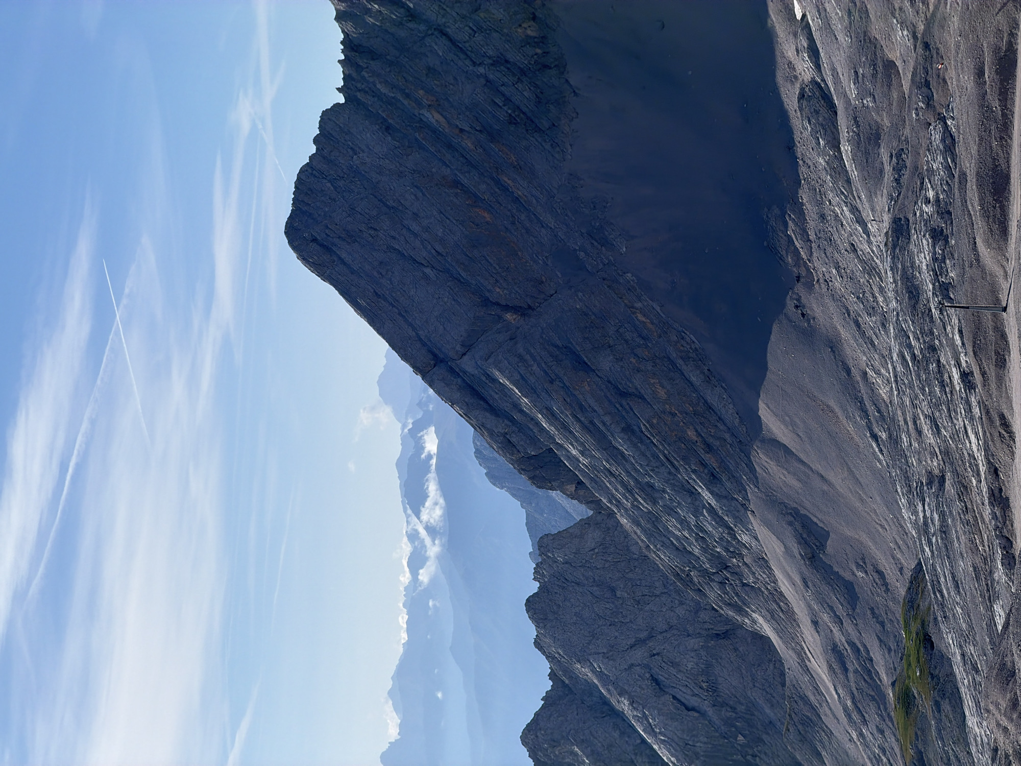 The image depicts a panoramic view of the Zugspitze, Germany's highest peak, located in Garmisch-Partenkirchen. The foreground is dominated by a rugged, rocky mountain slope that transitions into a vast, desolate landscape of scree and ice. In the mid-ground, a prominent, sharp-edged mountain peak rises dramatically, its textured surface indicative of steep inclines and geological strata. Further in the background, a series of progressively lighter, hazy mountain ranges recede into the distance, layered one behind another under a clear blue sky. The sky is adorned with wispy white clouds and two distinct contrails left by airplanes, forming a cross-like pattern. The overall scene conveys a sense of immense scale, natural grandeur, and the harsh beauty of a high-altitude alpine environment.