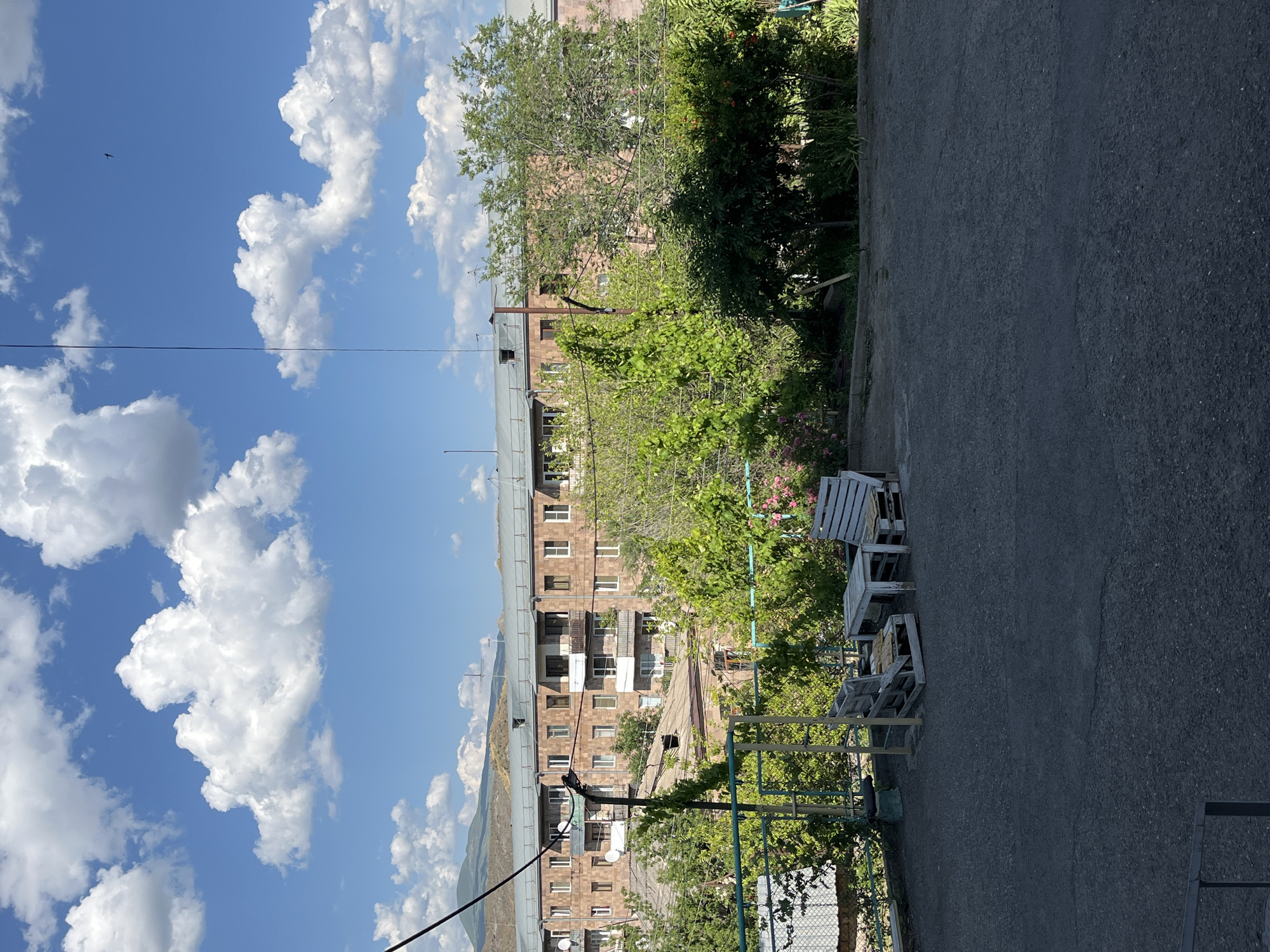The image depicts an outdoor scene in Yeghegnadzor, Armenia, on a bright, sunny day with scattered white clouds against a blue sky. In the foreground, there is a dark asphalt surface. Towards the center-left, a rustic outdoor seating area is visible, consisting of two chairs and a table constructed from wooden pallets. The chairs have light-colored cushions. Behind the seating area, a green metal fence runs horizontally. Beyond the fence, lush green trees and foliage dominate the middle ground, with some pink flowers also present. In the background, a multi-story apartment building with a reddish-brown brick facade and numerous windows is prominent. The building appears to be several stories high and has balconies. In the far distance, rolling hills and mountains can be seen, typical of the Armenian landscape. Power lines and a utility pole are also visible, intersecting the sky and landscape. A small bird is captured in flight in the upper right portion of the sky.