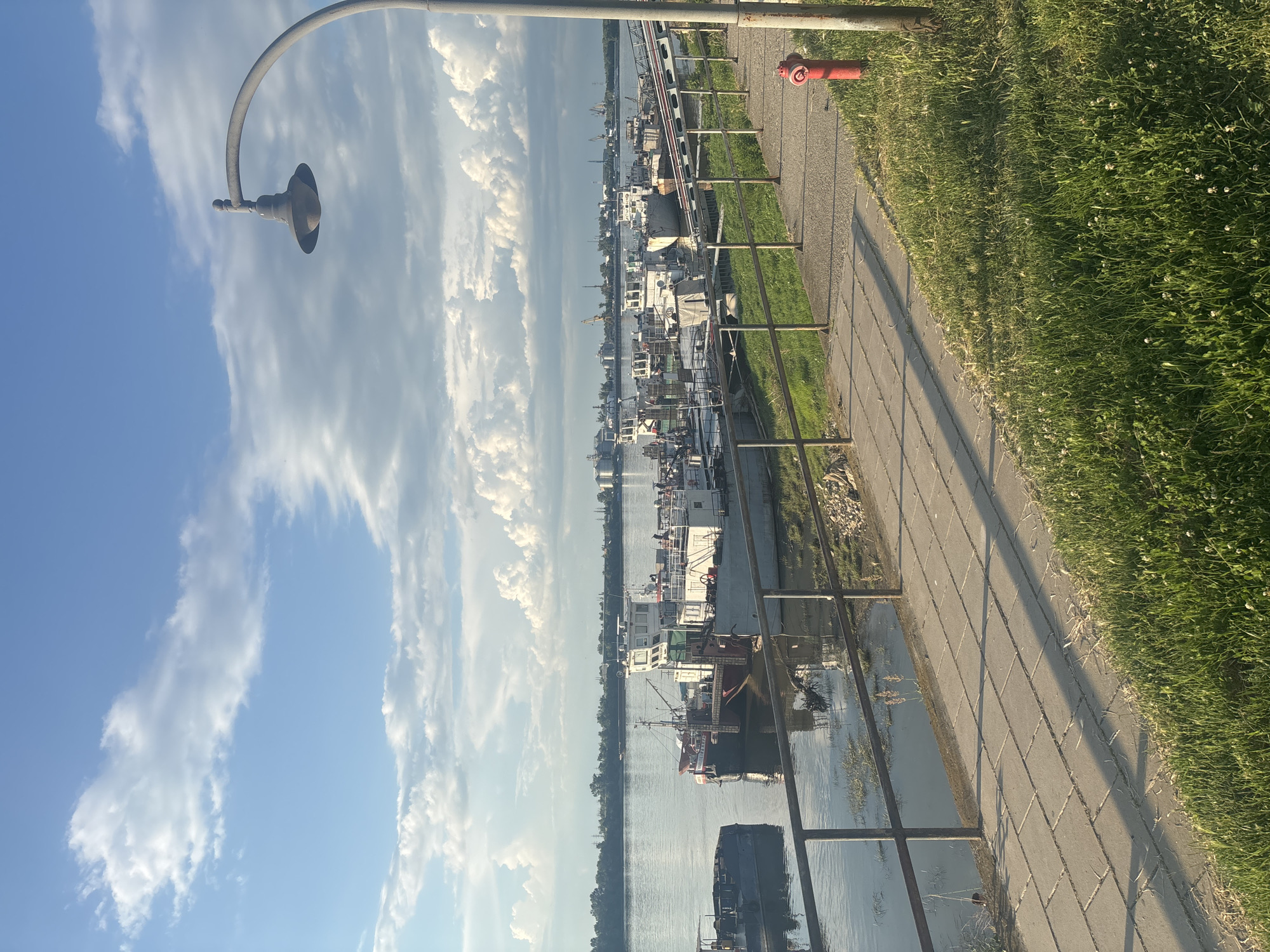 This is an eye-level, medium shot of a waterfront scene in Ruse, Bulgaria, captured during the daytime. The perspective is from a paved walkway adjacent to a river, with a metal railing separating the walkway from the water. Several barges and boats are moored in the river. The walkway is made of rectangular concrete pavers and is bordered by a grassy area on the right side of the frame. A tall, curved lamppost with an old-fashioned lamp is visible on the right, casting shadows on the walkway. A red fire hydrant is situated near the base of the lamppost. In the distance, across the river, is the Ruse industrial port, characterized by various port structures, cranes, and large industrial buildings. The sky above is a mix of blue with scattered white, puffy clouds. The overall atmosphere is peaceful and scenic, suggesting a calm day at the river.