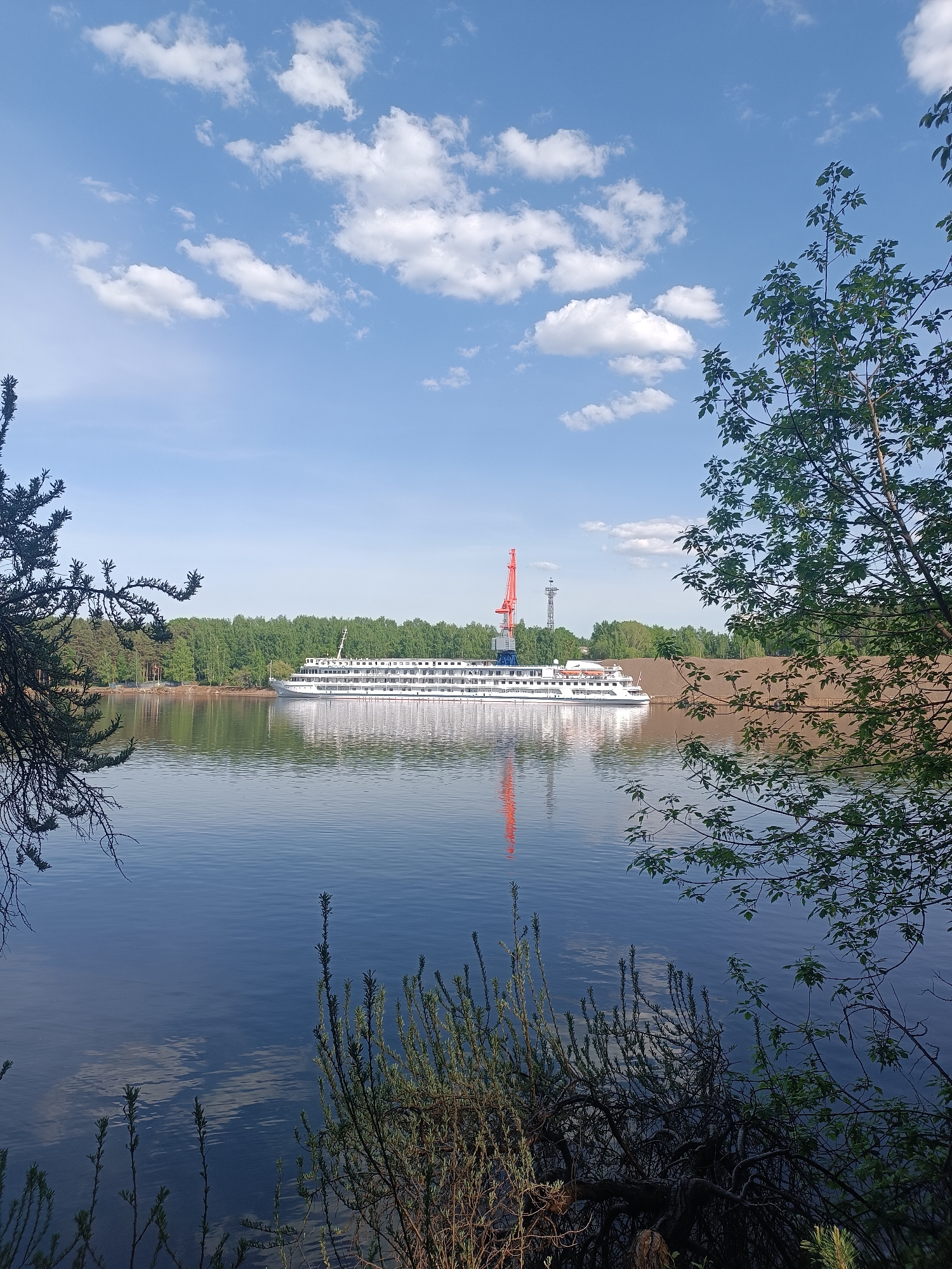 The image depicts a river scene in the city of Chaikovsky, Russia, with a large white cruise ship docked near the shore. In the background is a red port crane and a utility tower. The sky is blue with scattered white clouds. The river is calm, reflecting the ship and the crane. The foreground contains foliage, including tree branches and bushes on both sides. The riverbank is visible in the background, covered in green trees. There are no people visible.